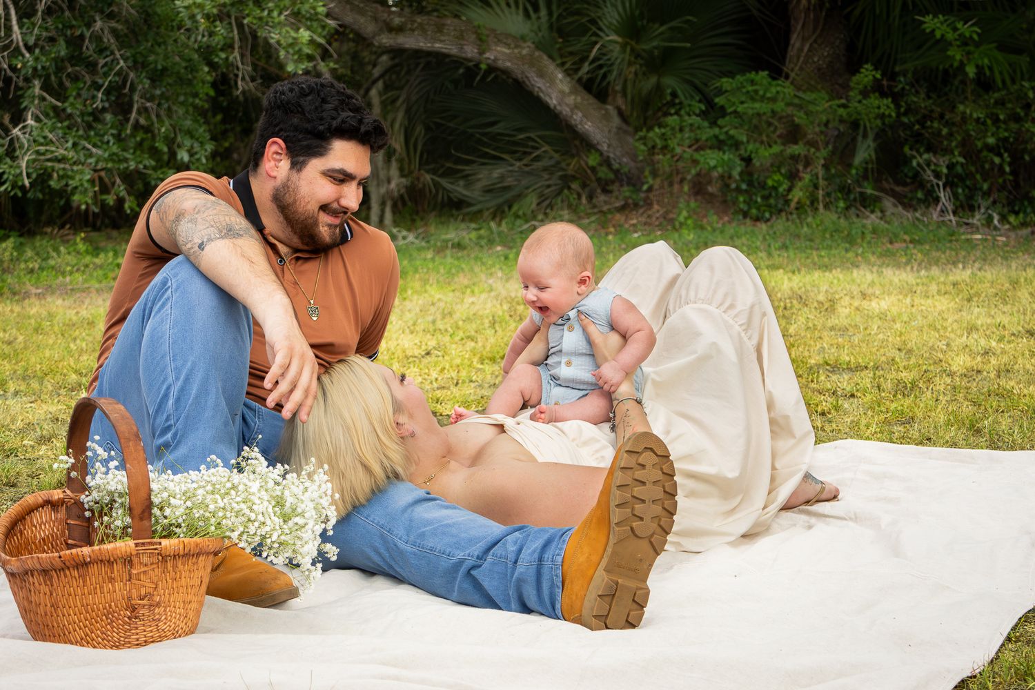 Young family enjoying a sunny picnic on grass, with father sitting and mother lying down holding baby.