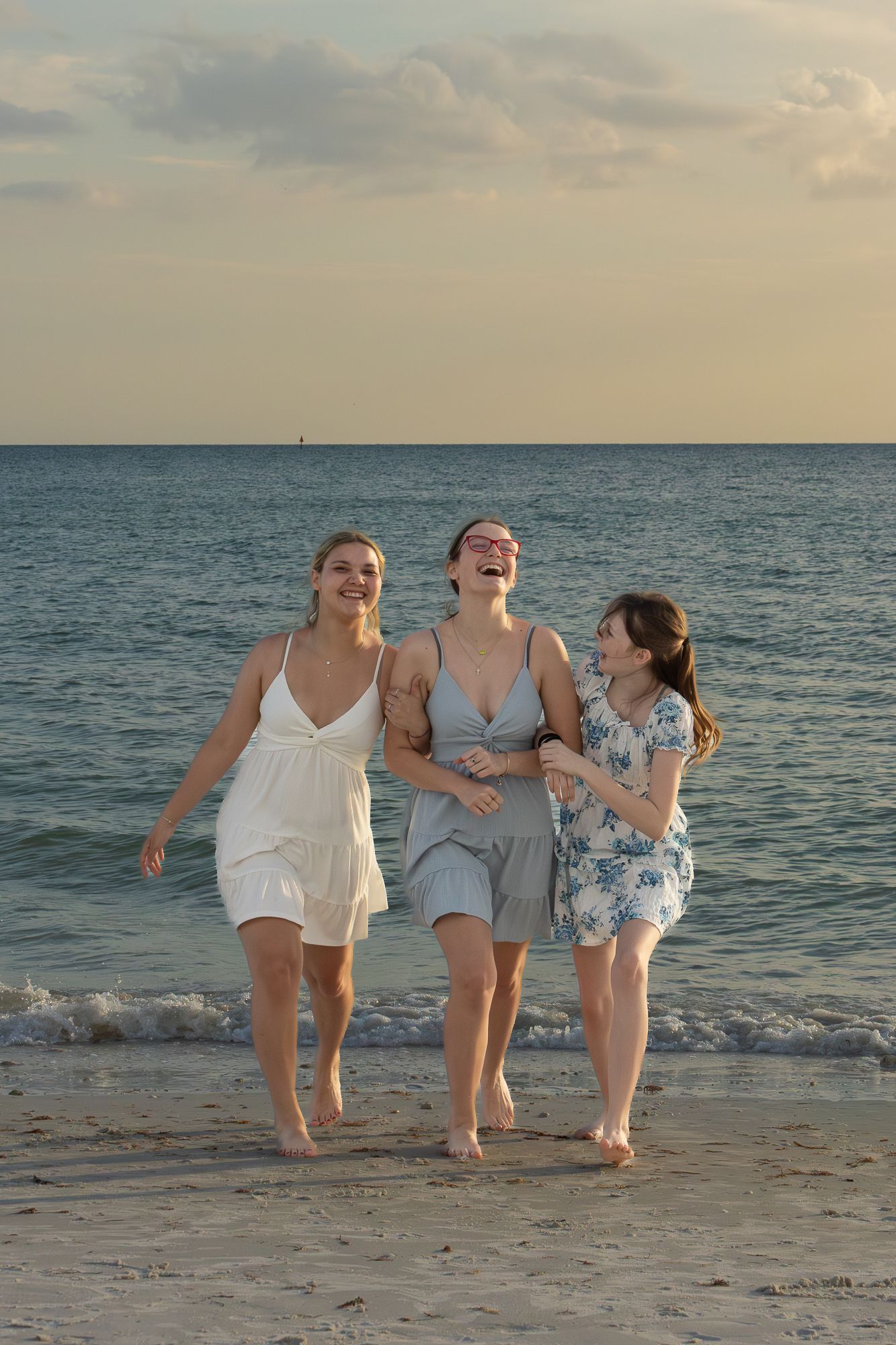 Three women laughing and walking barefoot along a sandy beach at sunset in summer dresses.