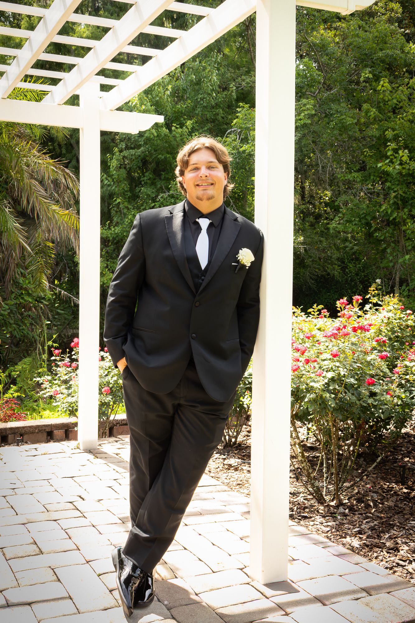 Groom in black tuxedo leaning against white pergola with rose garden backdrop.