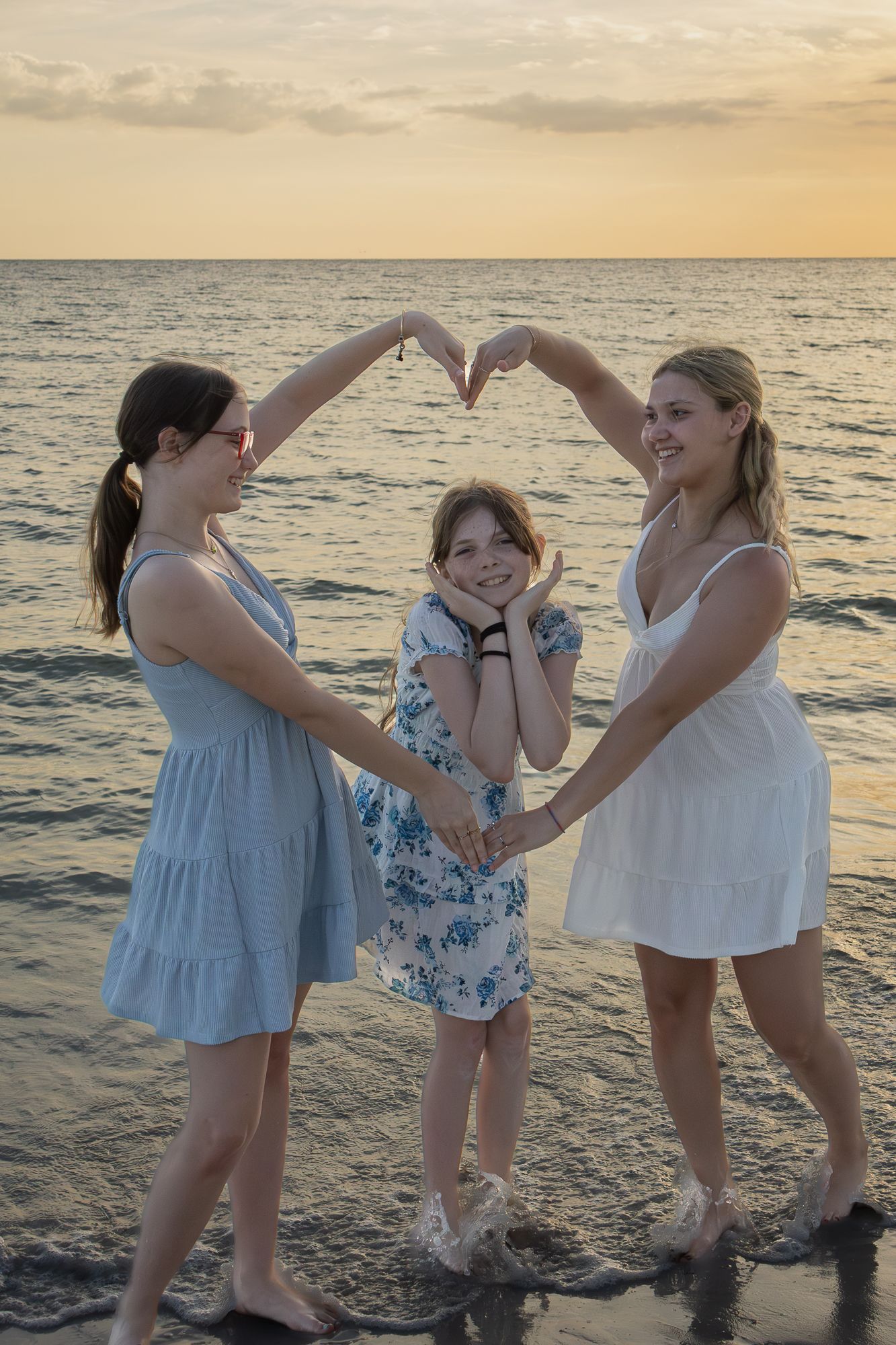 Two women forming a heart shape with their arms over a young girl on a beach at sunset.