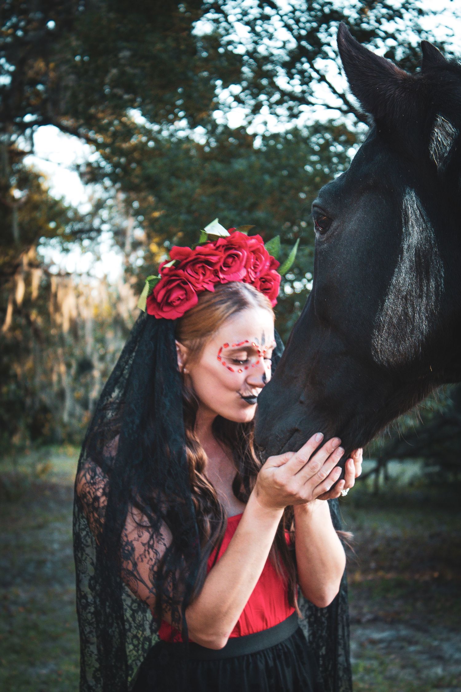 Woman in sugar skull makeup and red roses crown gently touching black horse.