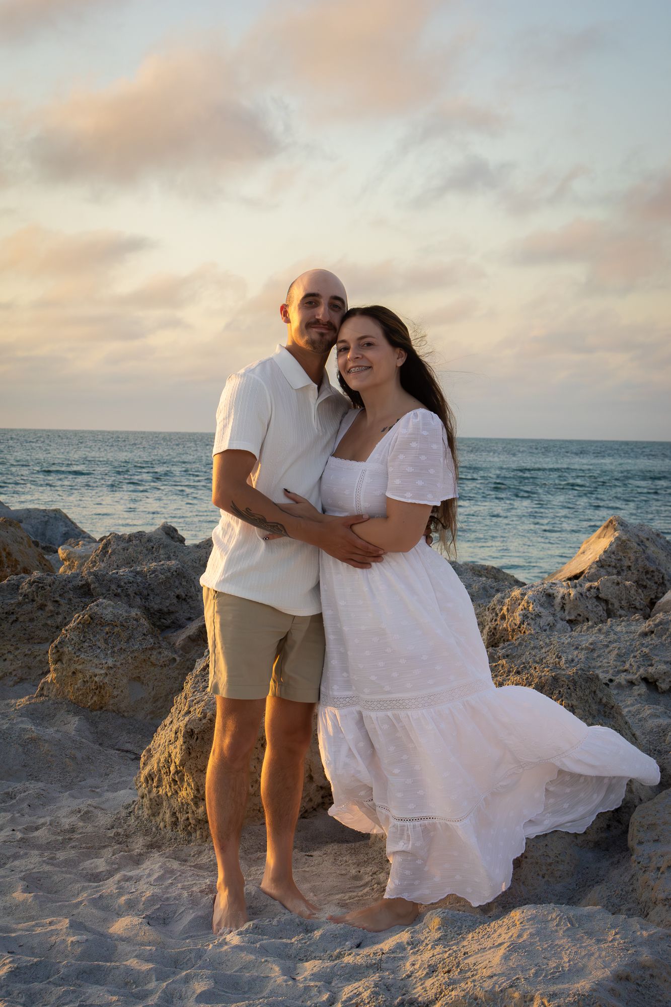 Couple embracing on a rocky beach at sunset, woman in white dress and man in white shirt and khaki shorts.