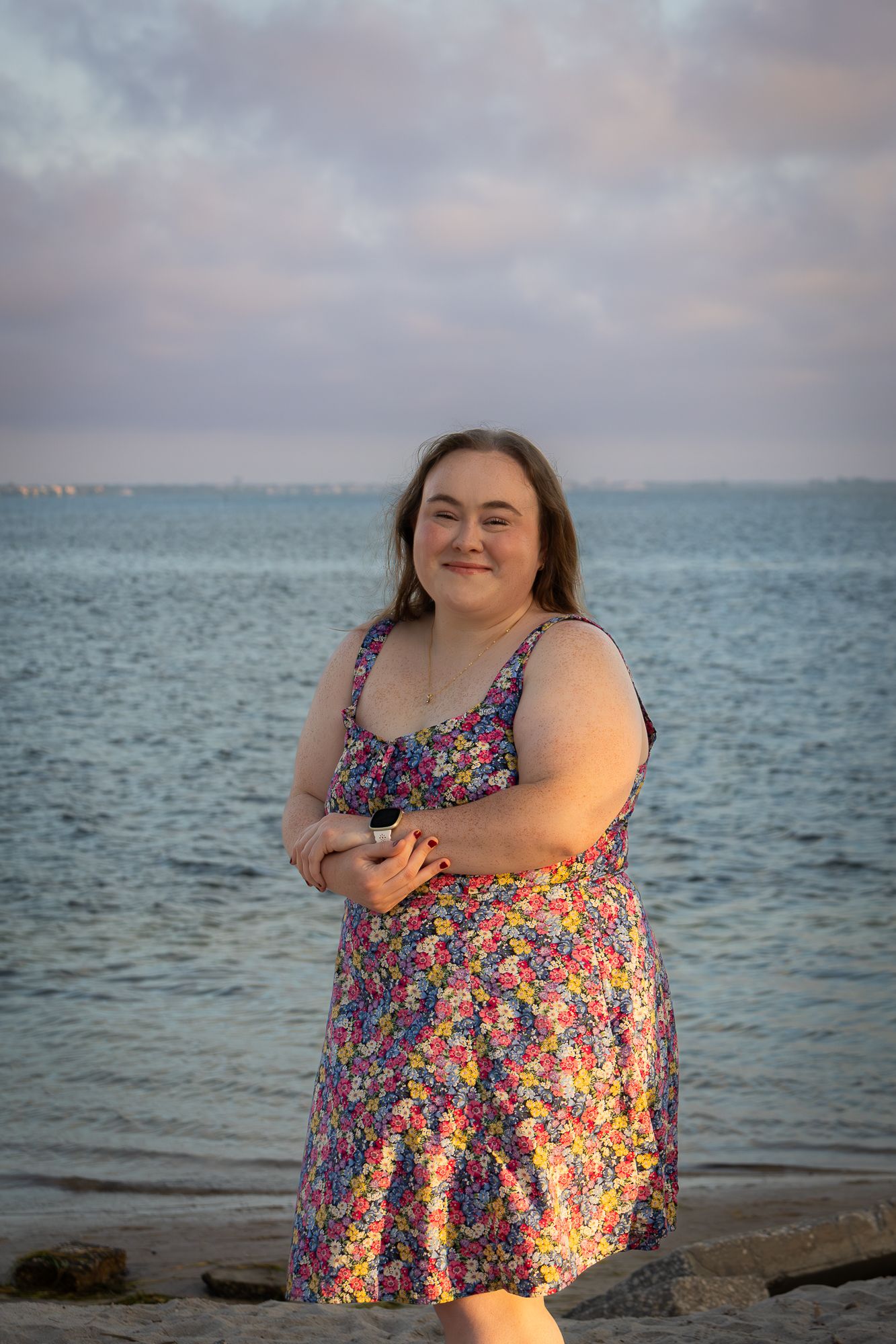 Happy woman in colorful floral sundress posing on rocky beach at sunset.