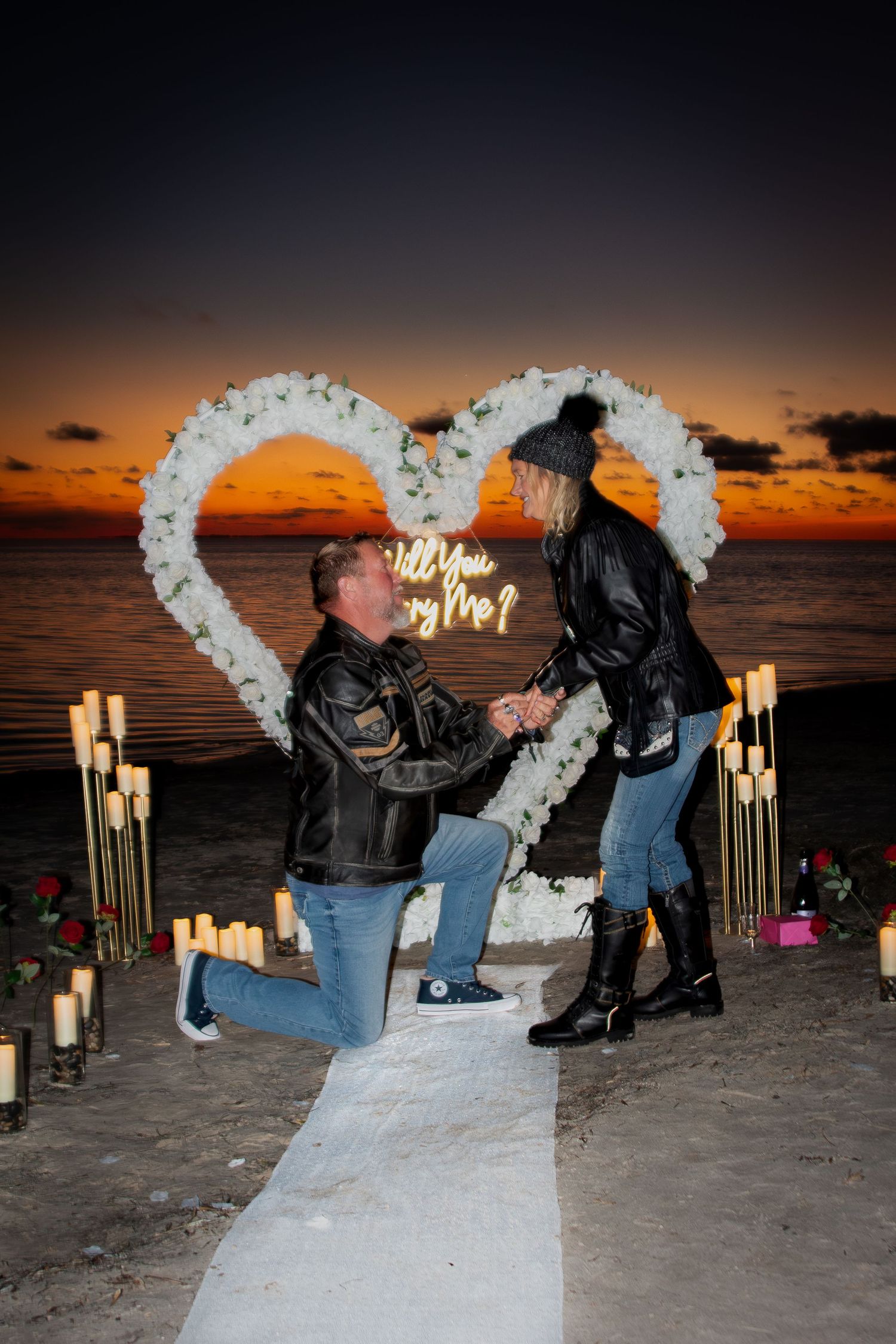 A man proposes on one knee to a woman at a romantic beach sunset setup with floral heart arch.