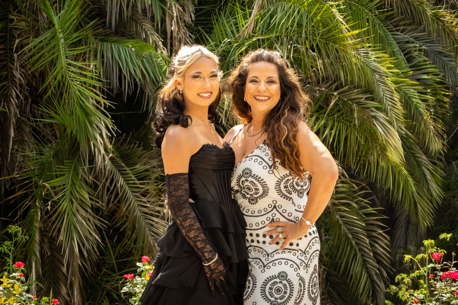 Two young women in elegant black and white formal prom dresses posing before lush tropical palm trees.