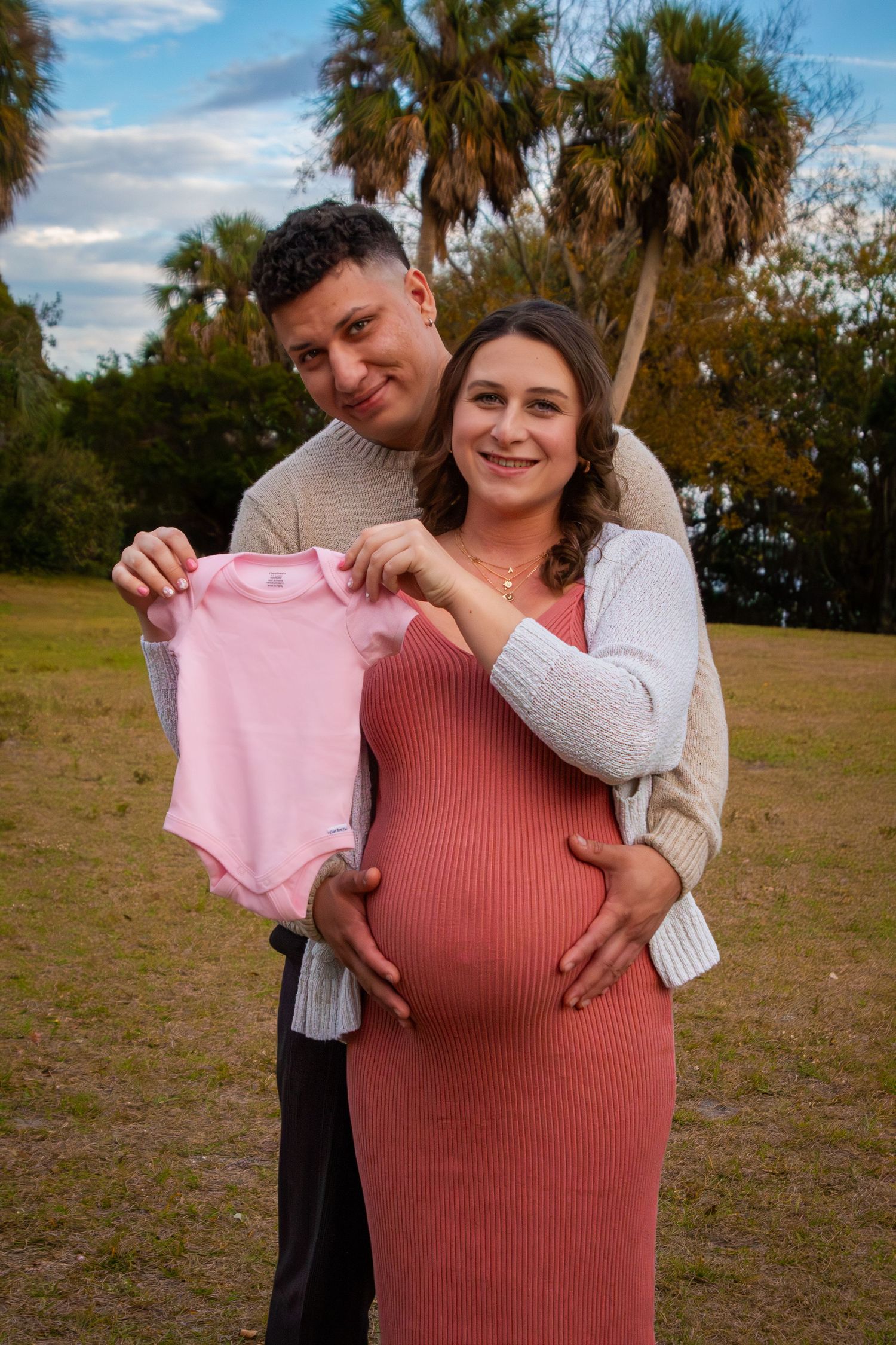 Couple holding pink baby onesie outdoors, announcing baby girl pregnancy.