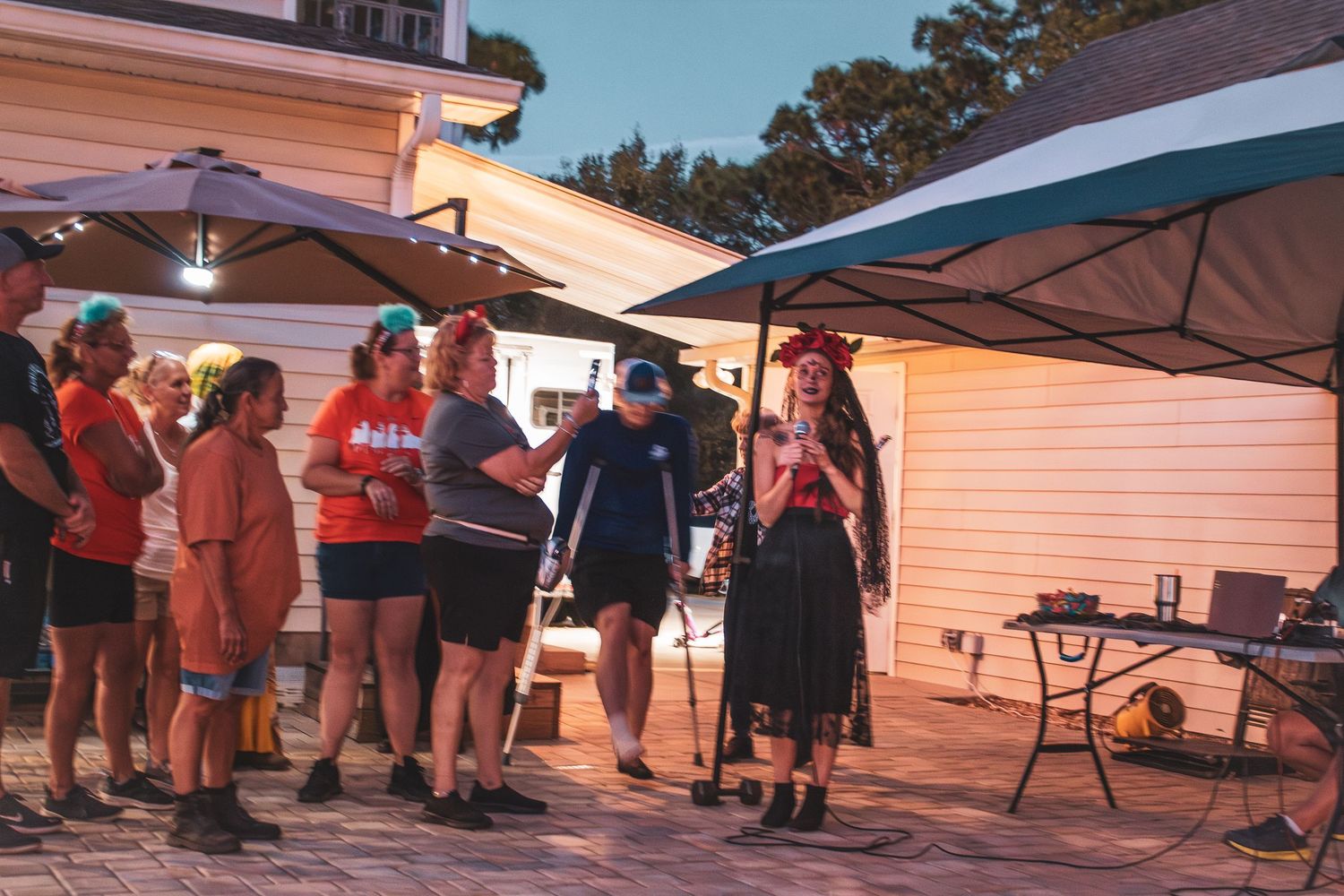 A group of people gather under outdoor canopy tents at a backyard evening event near a house.