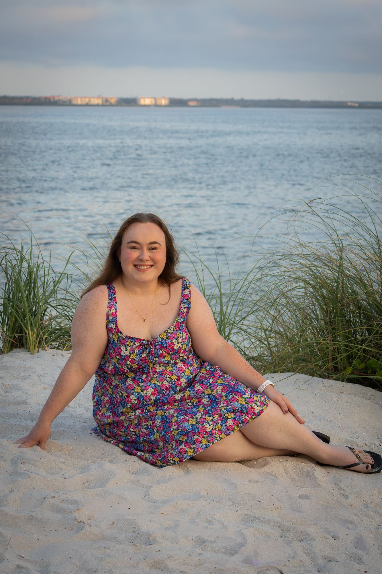 Smiling woman in floral dress sitting on white sand beach at dusk.