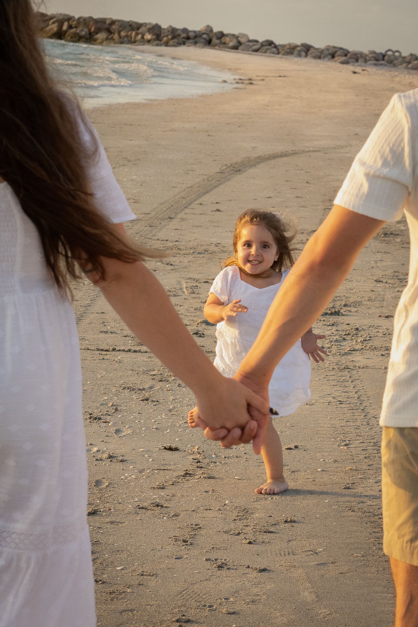 Smiling little girl in white dress running toward parents holding hands on a sunny beach.