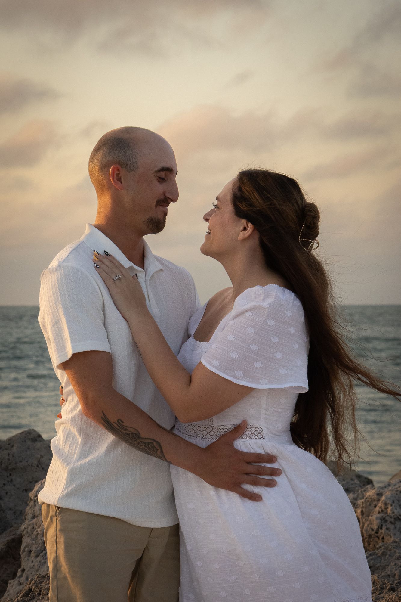 A loving couple gazes at each other on a rocky shoreline at sunset, both dressed in white and cream tones.