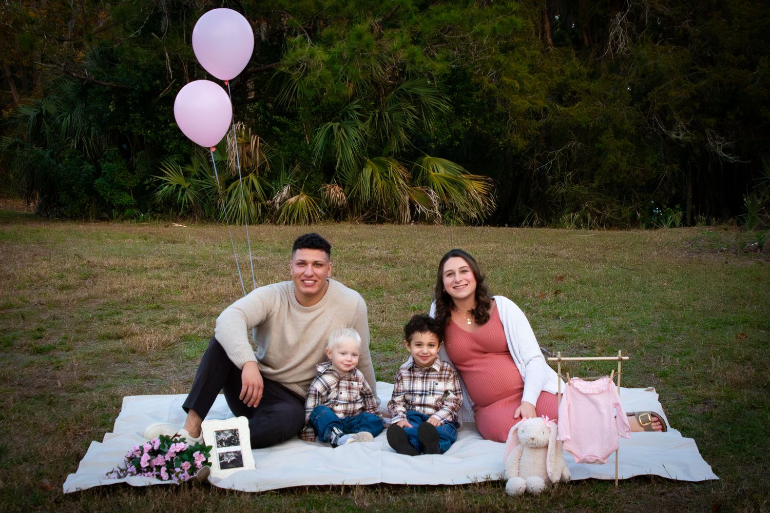 Family maternity photo with couple and two young boys on a blanket with pink balloons.