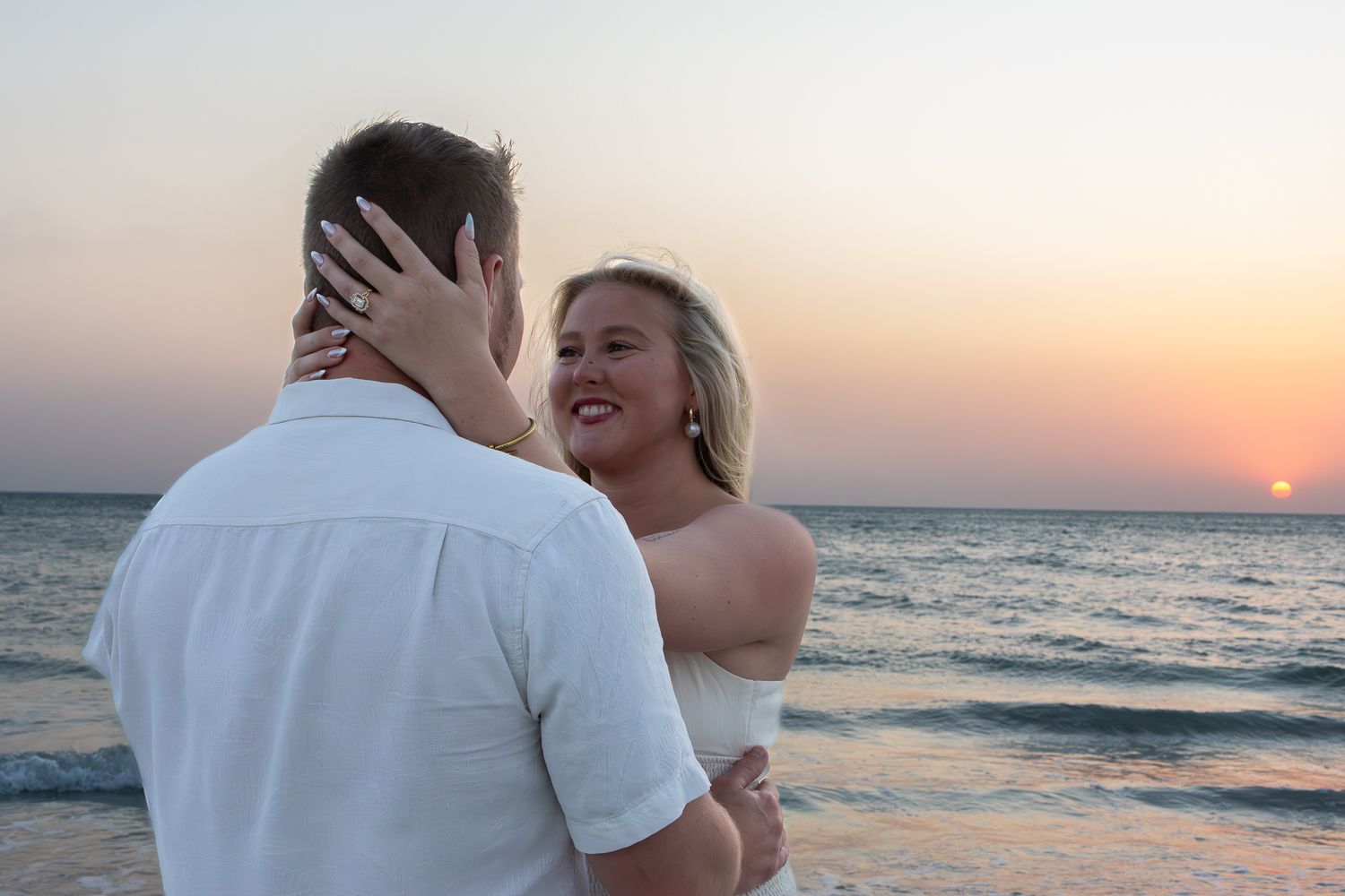 Couple embracing on beach at sunset, woman smiling joyfully with hand on mans face.