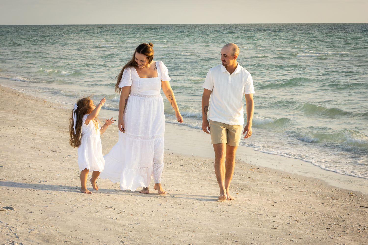 Family of three holding hands and walking along a white sandy beach near the ocean shoreline.