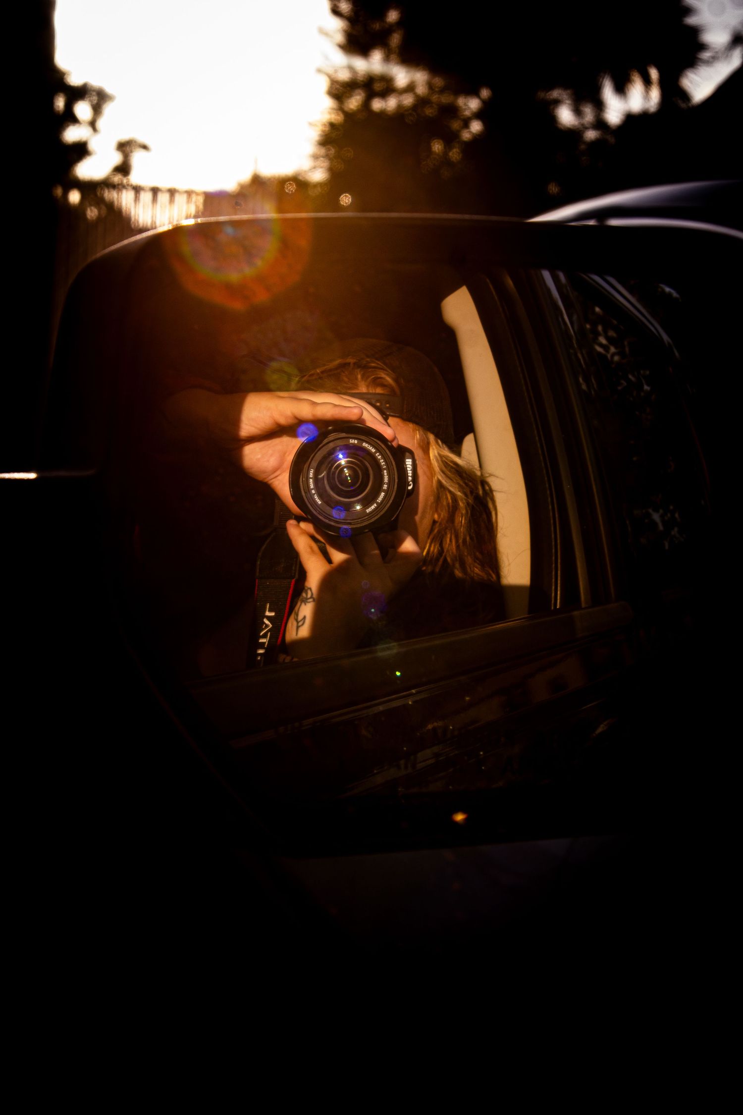 Photographer capturing self-portrait in car side mirror during golden hour sunset with warm lens flare.
