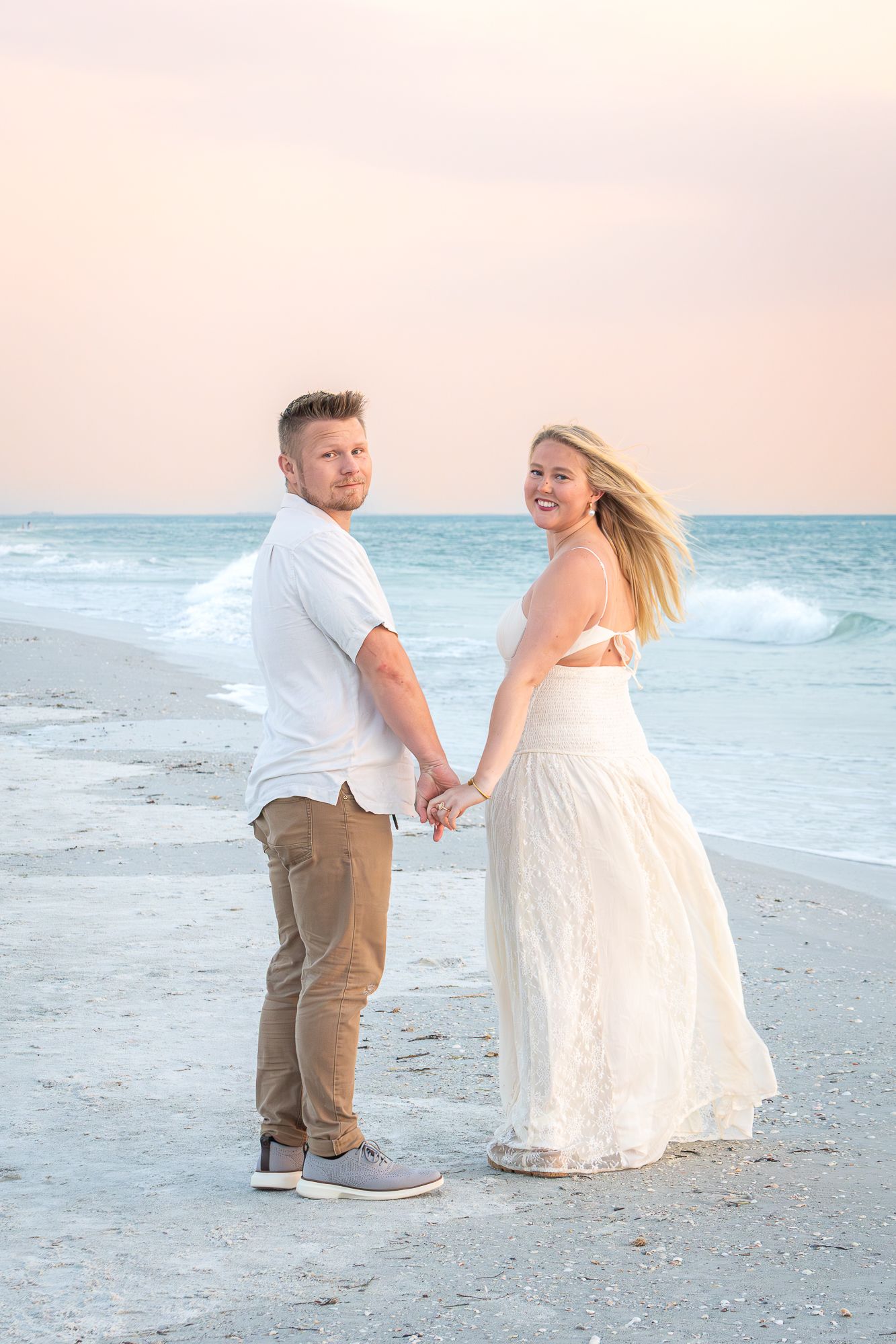 Happy engaged couple holding hands on a beach at sunset, smiling back at the camera.