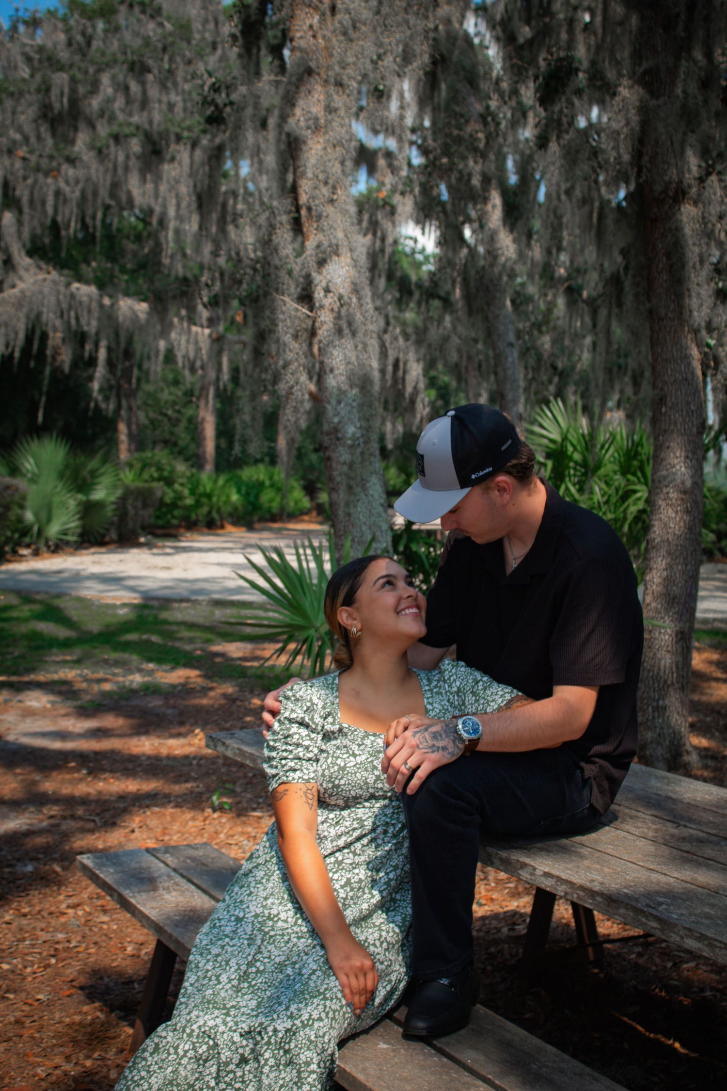 Romantic couple sharing an intimate moment seated on wooden bench under Spanish moss-draped trees in park.