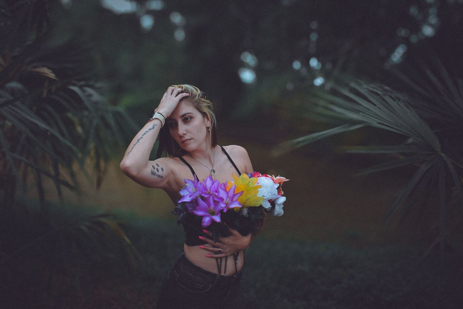 Young woman with tattoos holding colorful flowers bouquet poses among lush tropical palm leaves in moody outdoor setting.