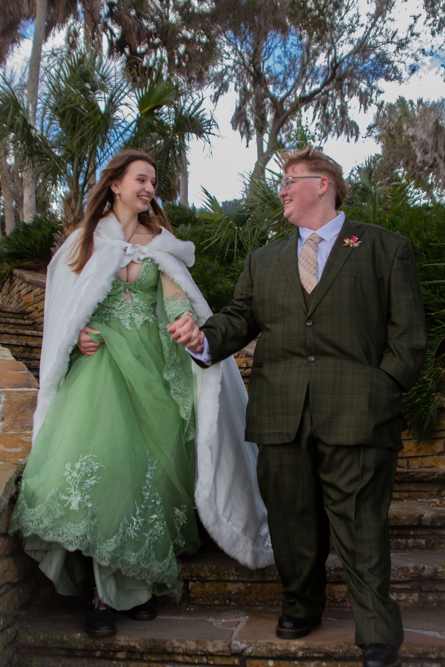 Bride in green lace wedding gown with white cape walking with groom in tweed suit outdoors.
