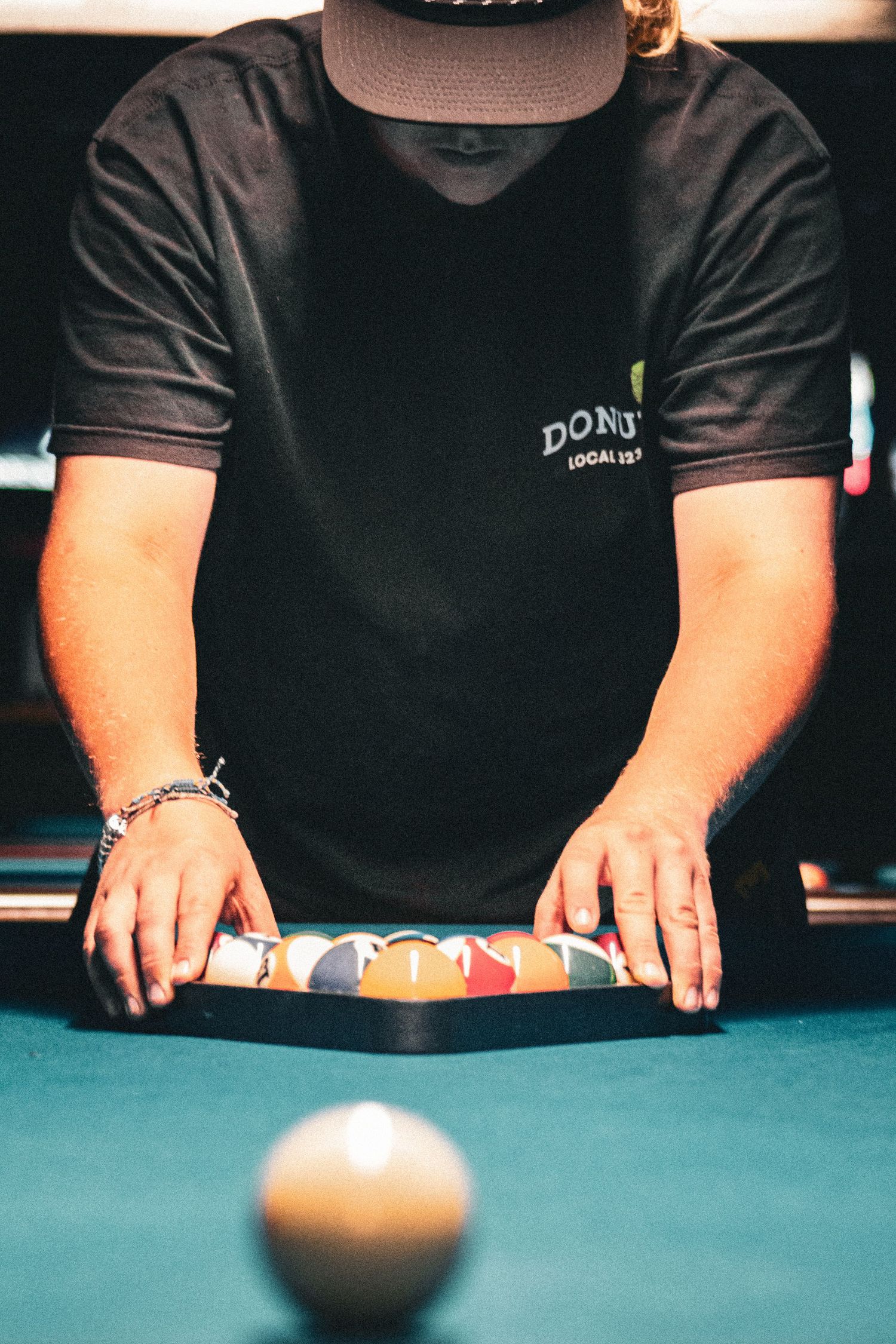 Man in black Donut shirt racking billiard balls on a green pool table, cue ball in foreground.