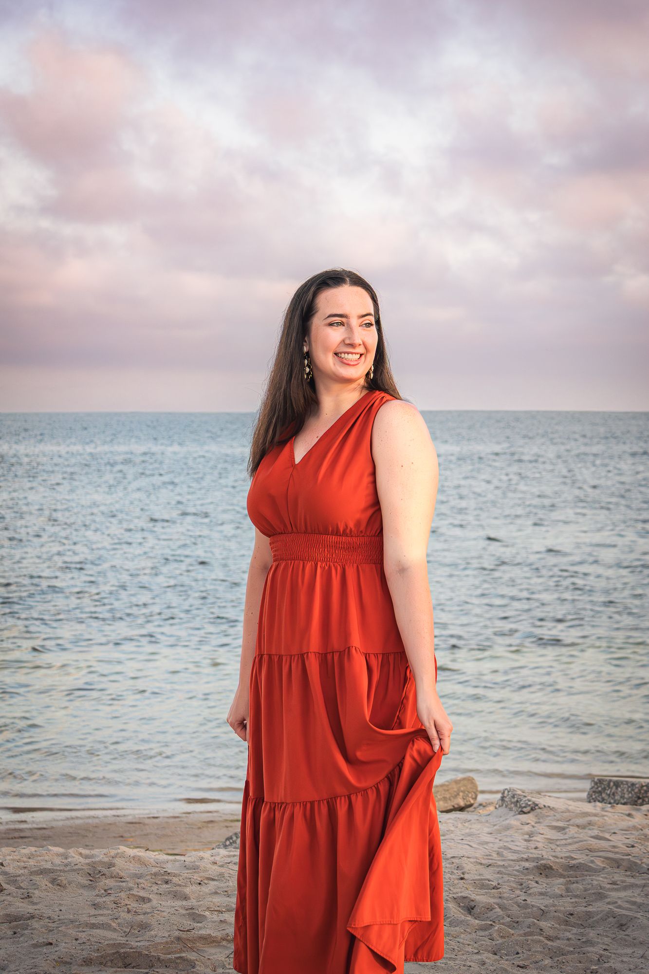 Woman in red maxi dress standing on beach at sunset with ocean backdrop.