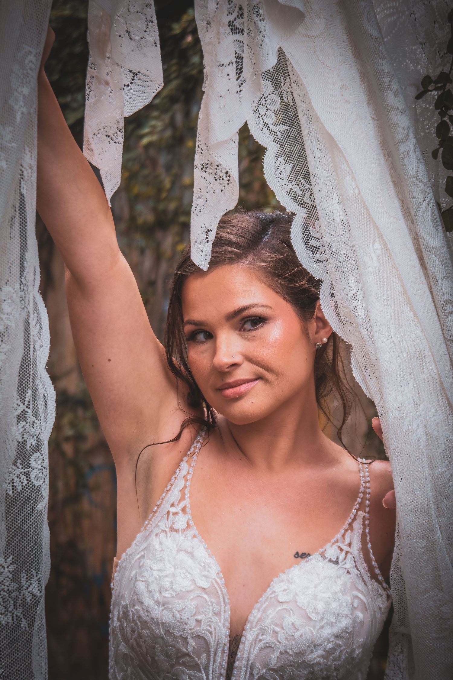 Beautiful bride in lace wedding gown holding white fabric with arm raised in outdoor setting.