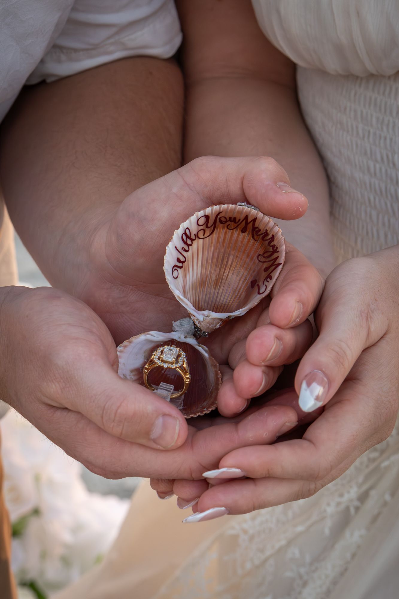 Couple holding a seashell ring box with an engagement ring and Will You Marry Me written inside at a beach proposal.