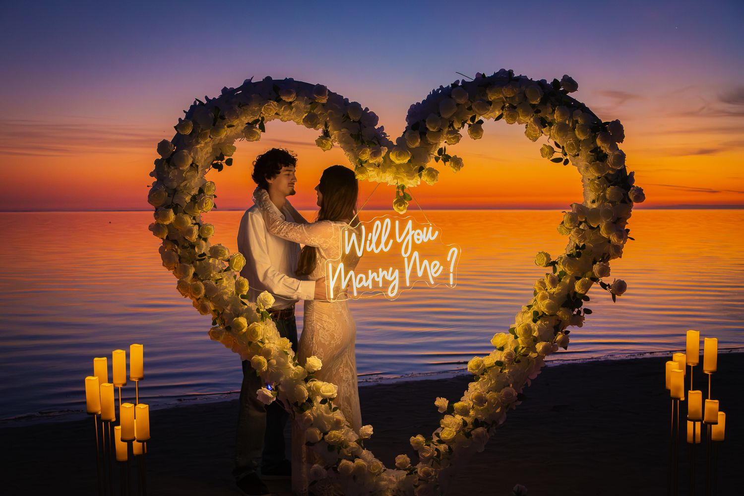 Romantic beach proposal setup with a floral heart arch, Will You Marry Me neon sign, and candles at sunset.