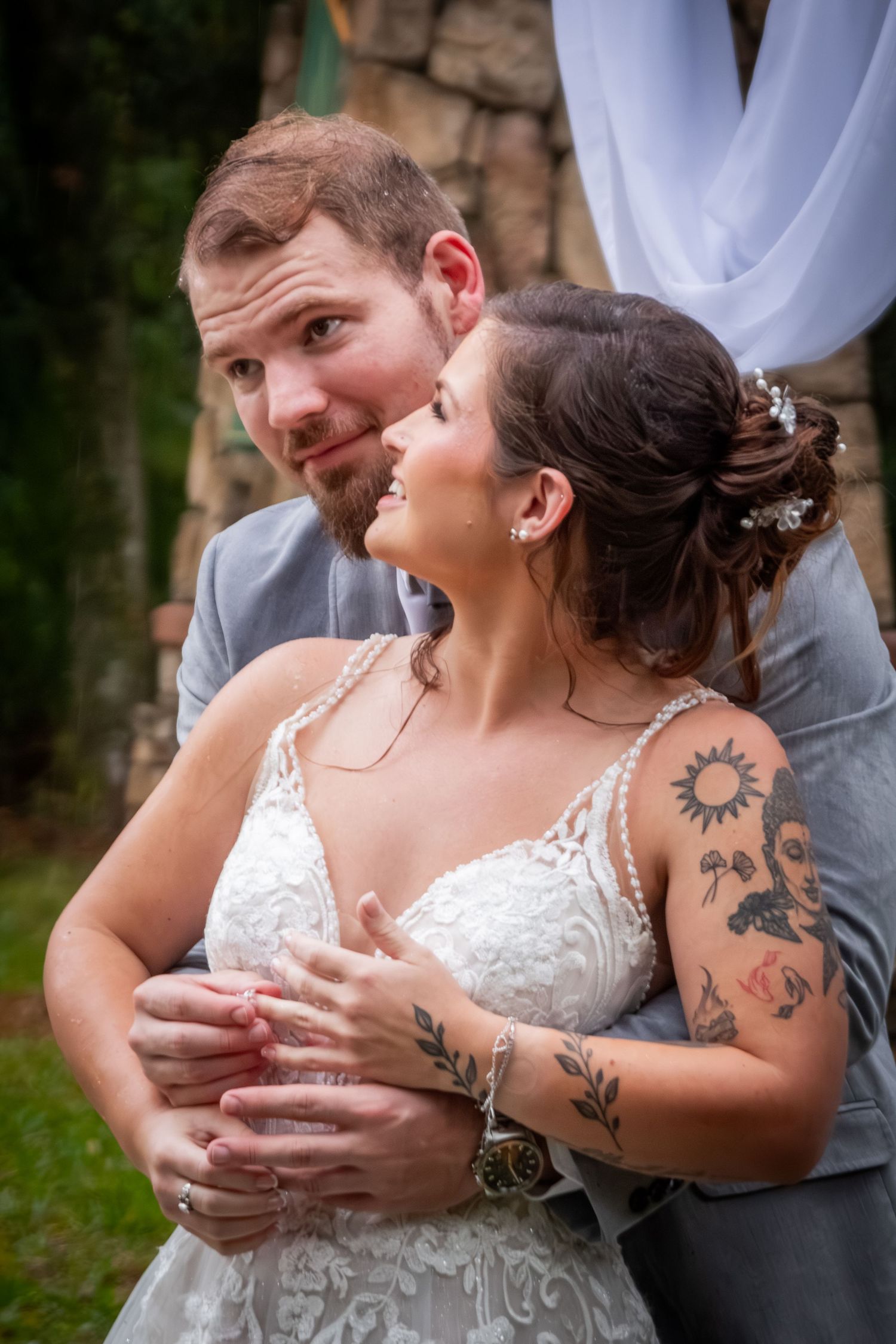 Groom in gray suit embracing tattooed bride in lace wedding dress during outdoor ceremony.