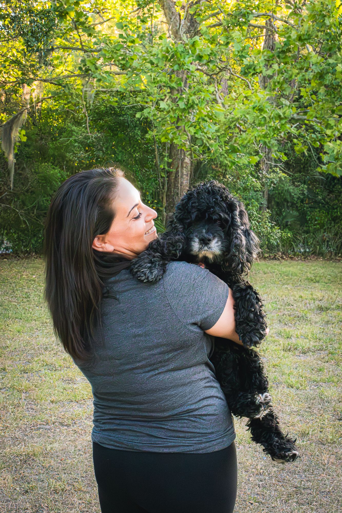 Woman in gray shirt holding a black curly-haired dog while standing in a lush green backyard.
