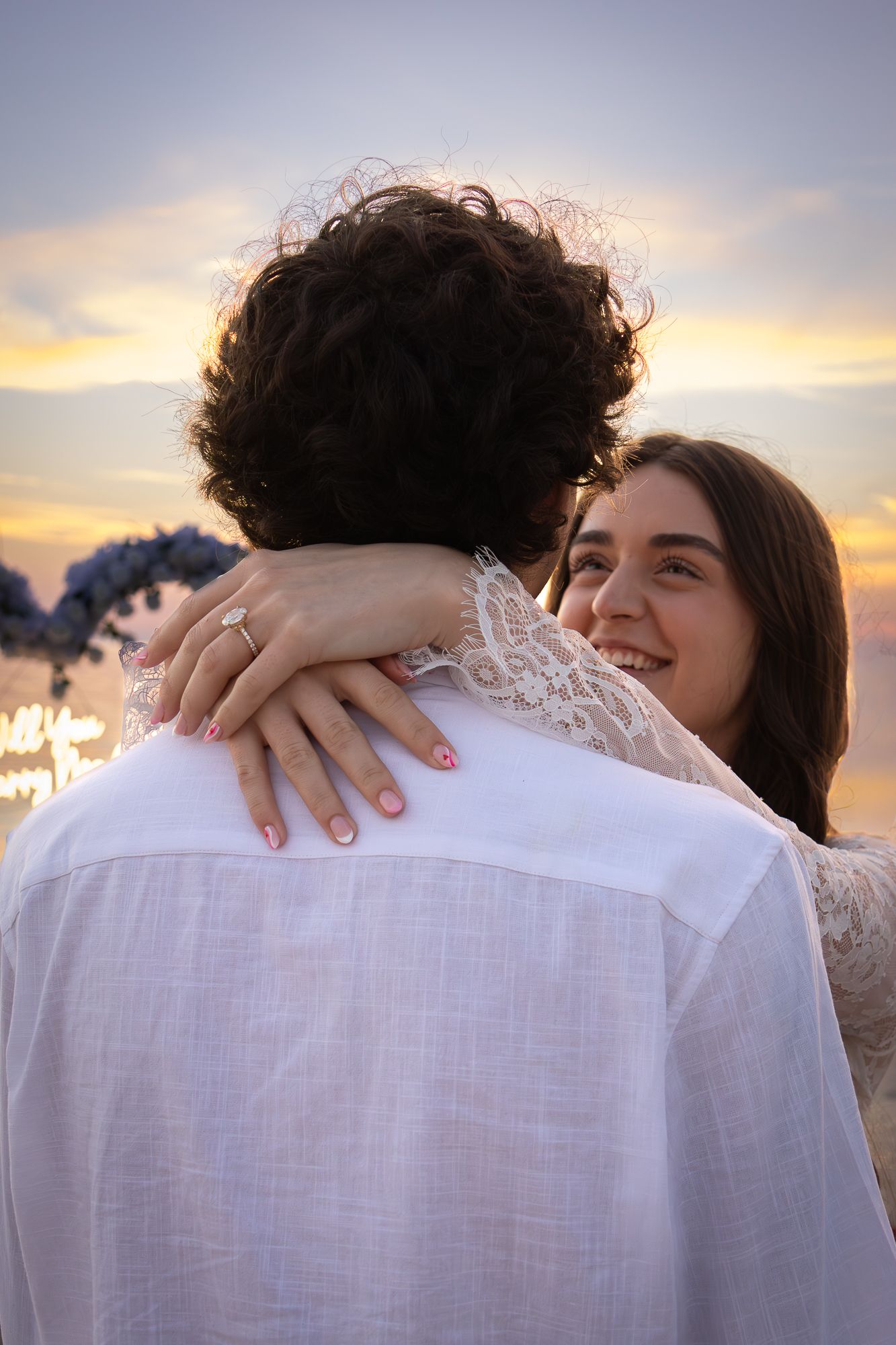 Couple embracing at sunset, woman smiling with engagement ring visible on hand resting on mans shoulder.