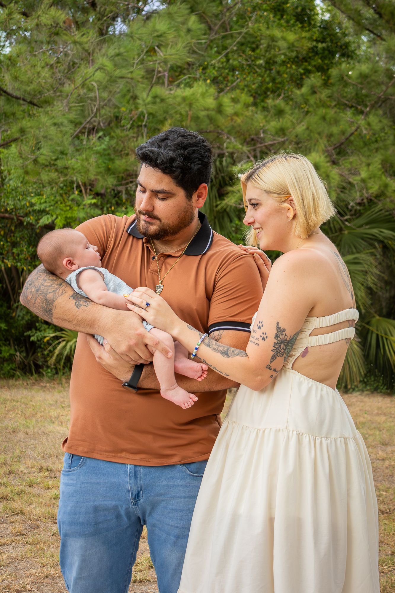 Tattooed couple in casual attire cradling newborn baby outdoors in nature.