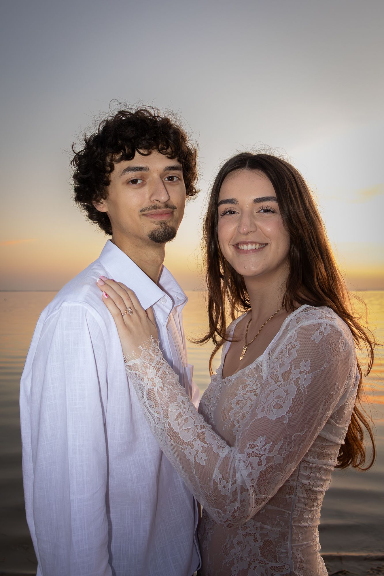 A smiling couple poses together at sunset by the ocean, the woman in a lace dress embracing the man in white.