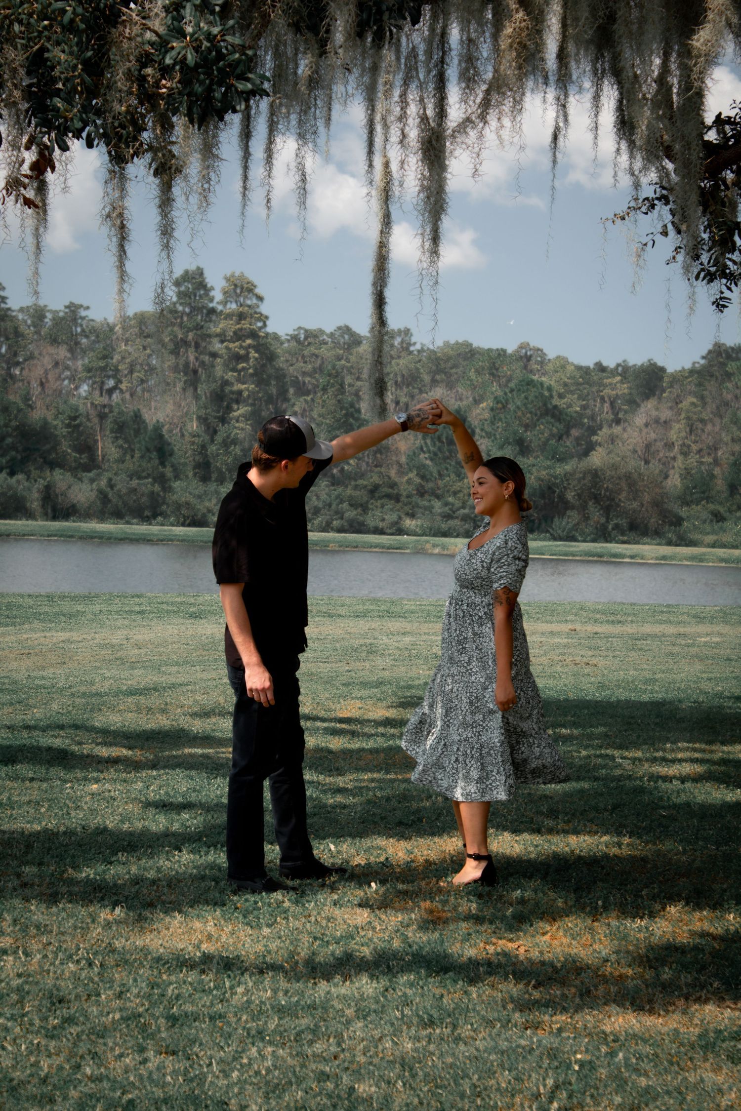 Couple dancing together on green lawn near lake under hanging Spanish moss trees on sunny day.