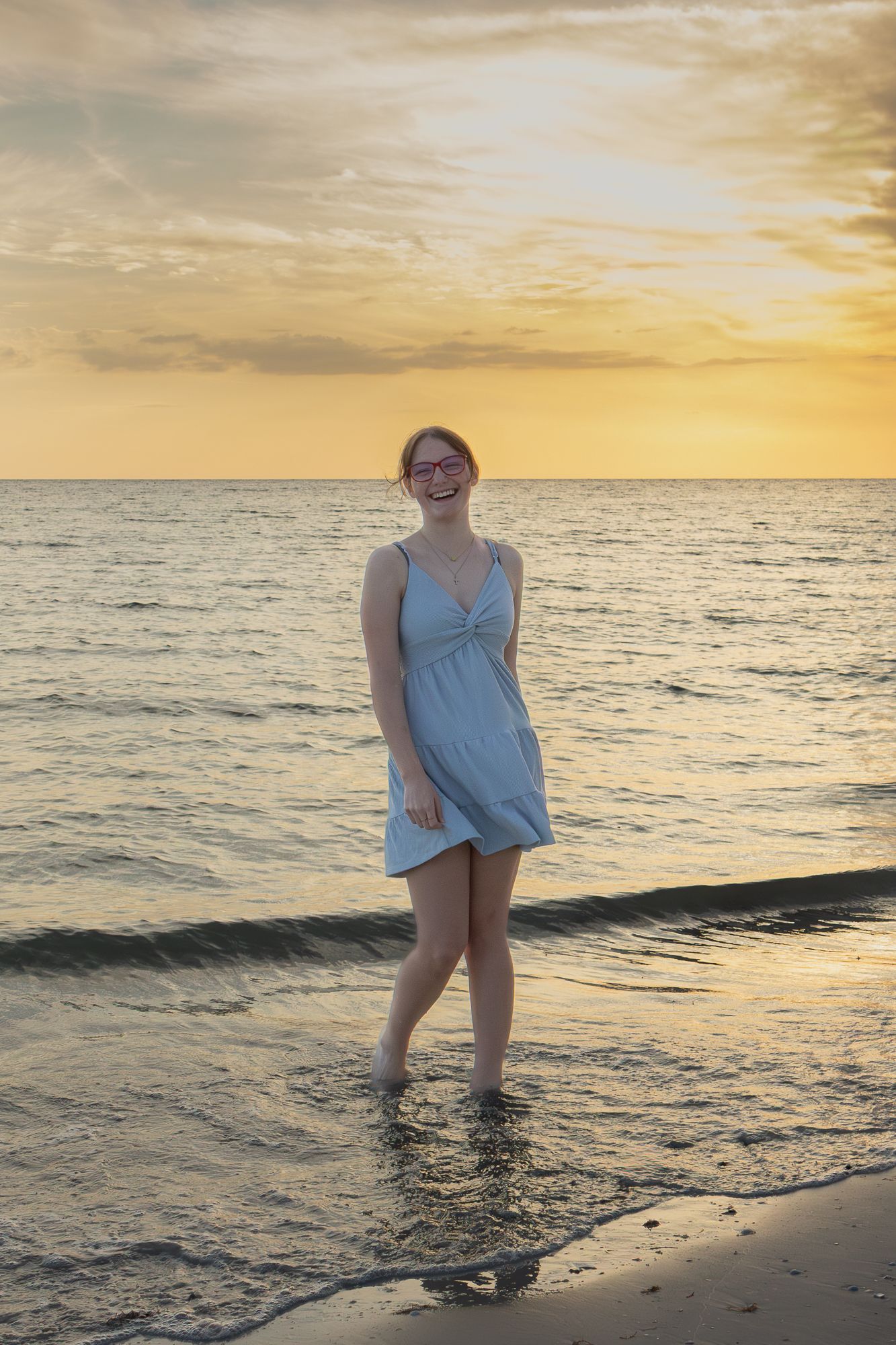 Young woman in a light blue dress standing barefoot in ocean waves during a golden sunset.