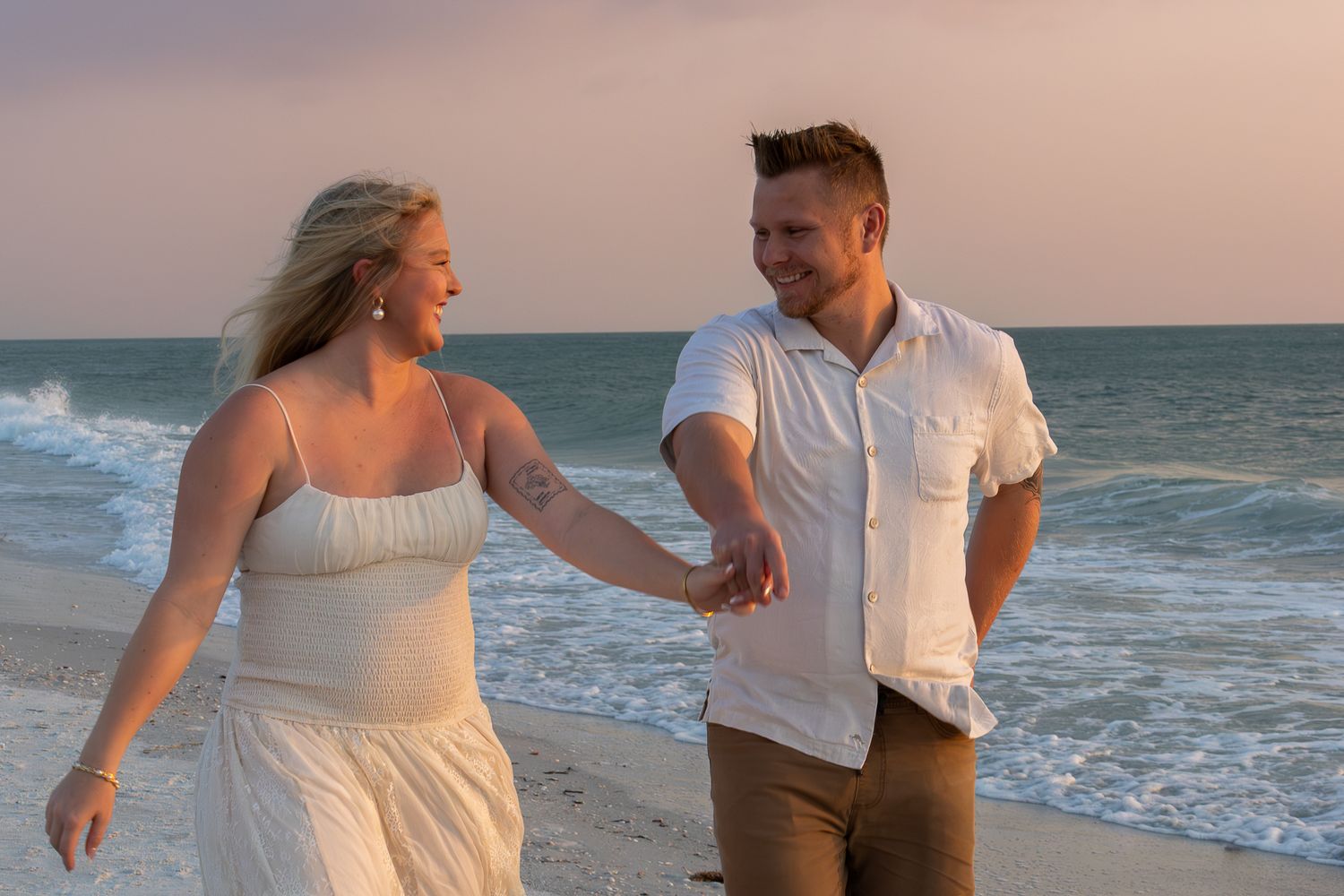 Smiling couple holding hands and walking on a sandy beach during a colorful sunset.
