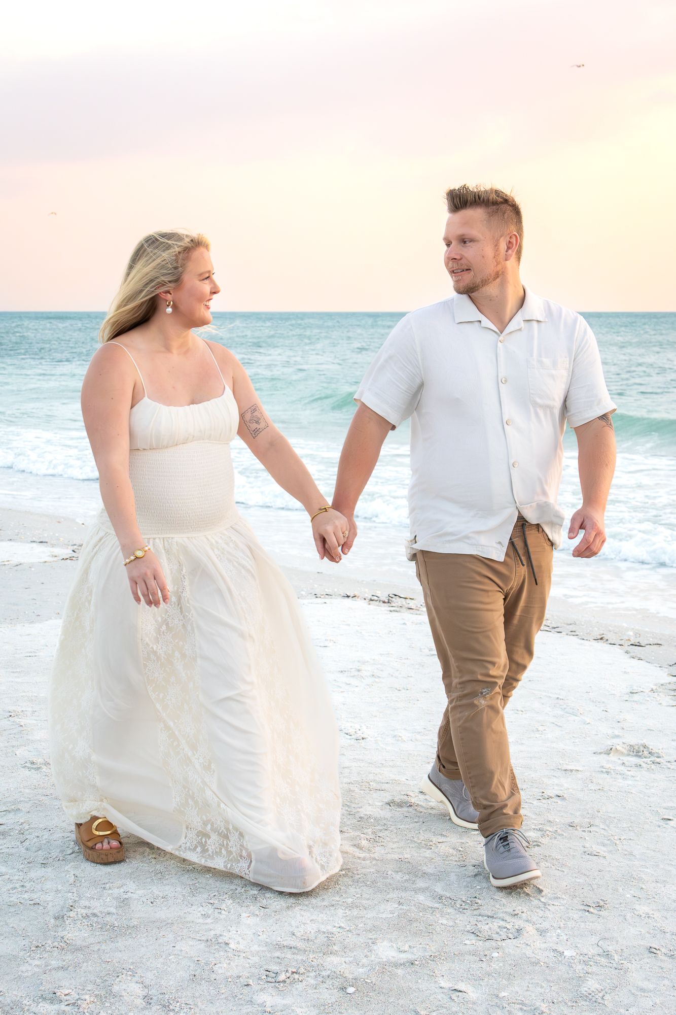 A smiling couple walks hand in hand on a beach at sunset, the woman in a white gown and the man in khaki pants.
