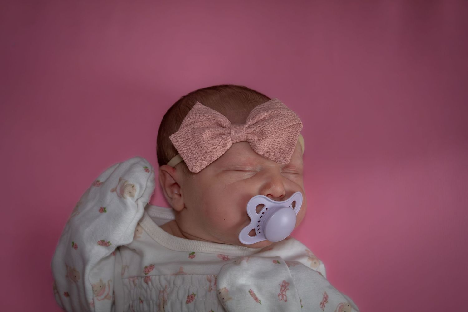 Newborn baby with pink bow headband and pacifier against a pink background.