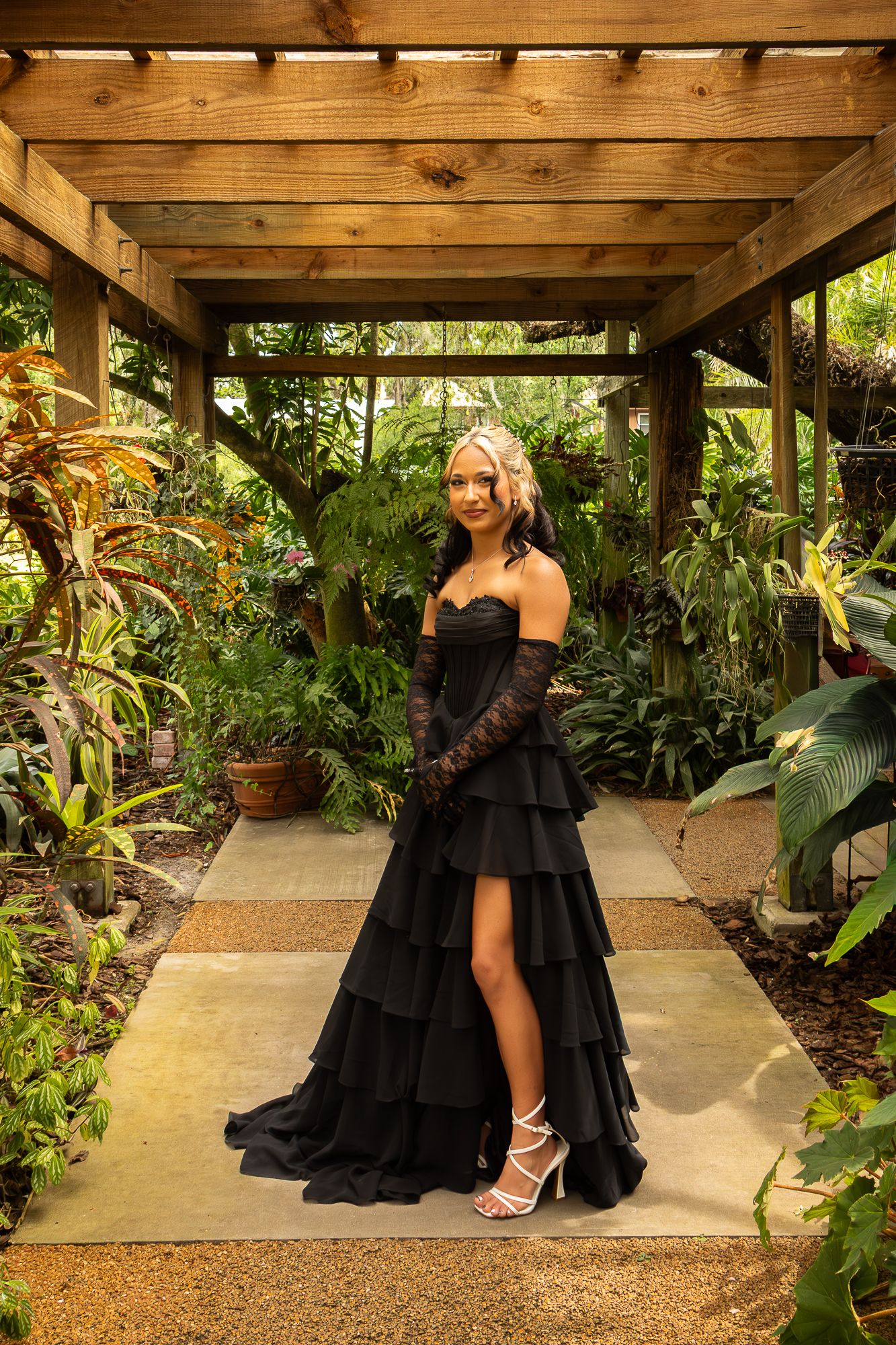 Elegant woman in black off-shoulder tiered gown with lace gloves posing in tropical garden.