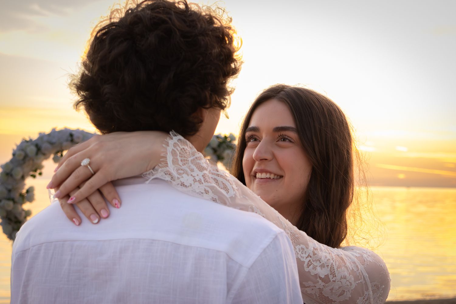 Happy bride with engagement ring embracing groom at golden sunset beach backdrop.
