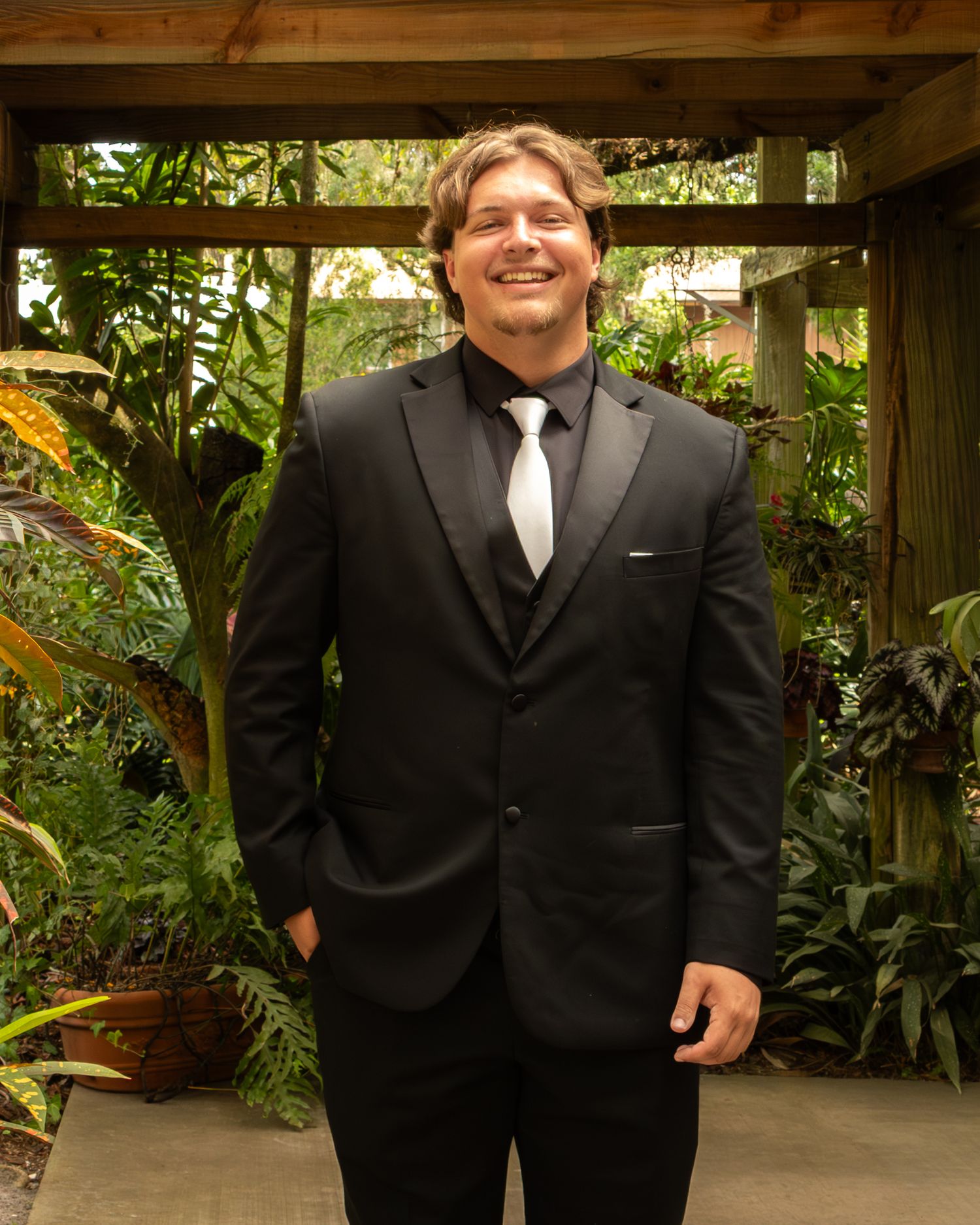 Man in black tuxedo smiling in lush garden setting.