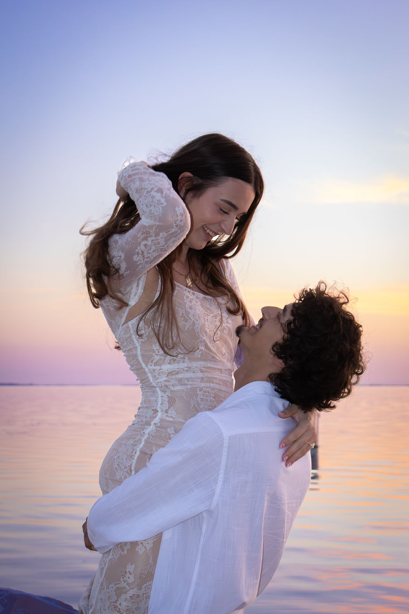 A joyful couple shares a romantic moment at sunset by the ocean, the man lifting the woman in a white lace dress.