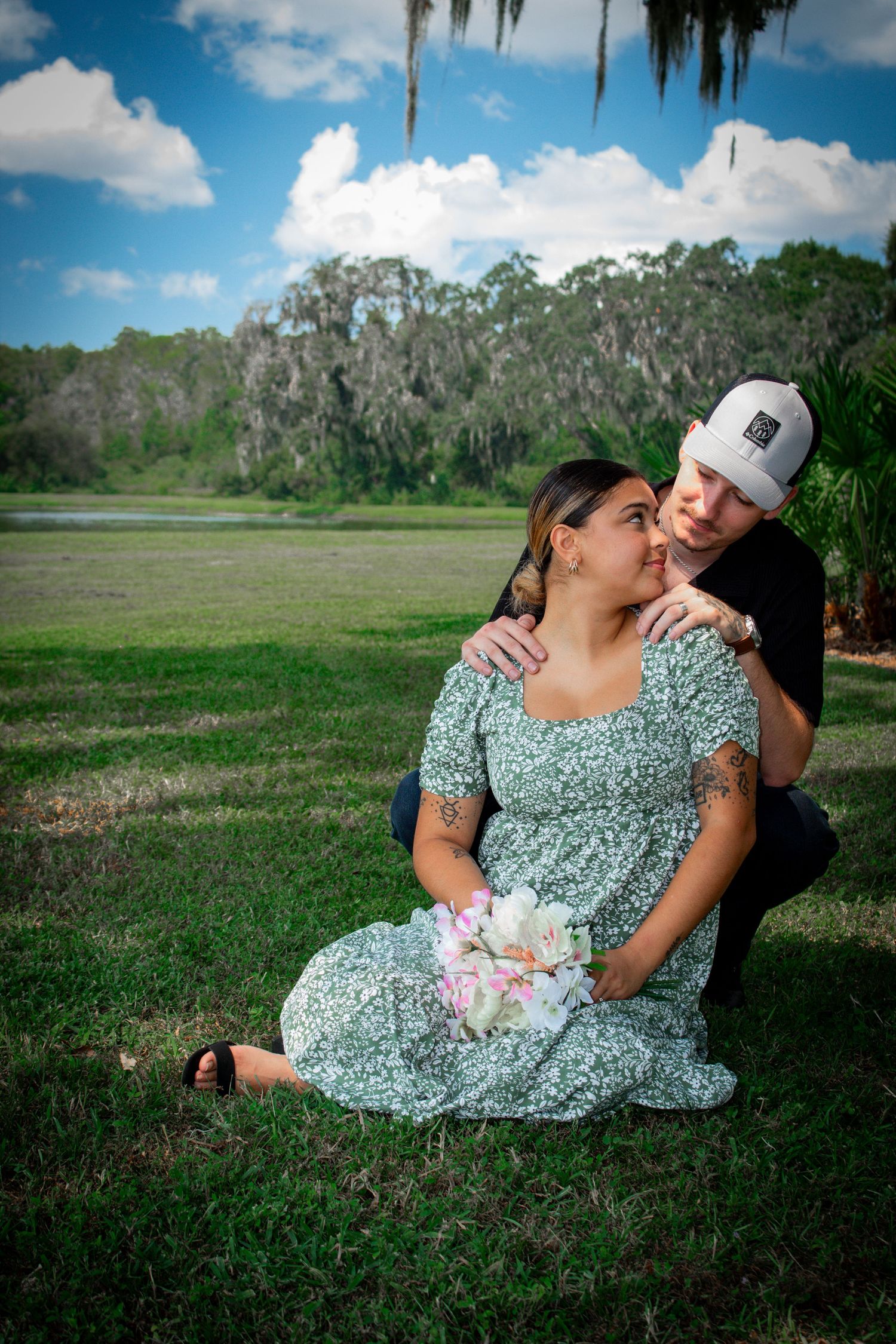 Couple sitting on grass outdoors, woman in floral dress holding flowers, man in white cap embracing her, lush green park backdrop.