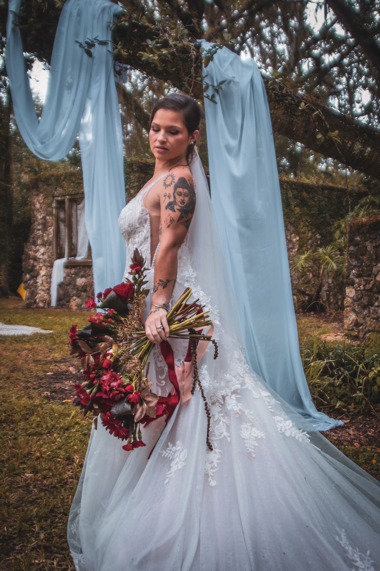 Tattooed bride in white lace gown holds red bouquet beneath blue draped fabric at outdoor wedding.