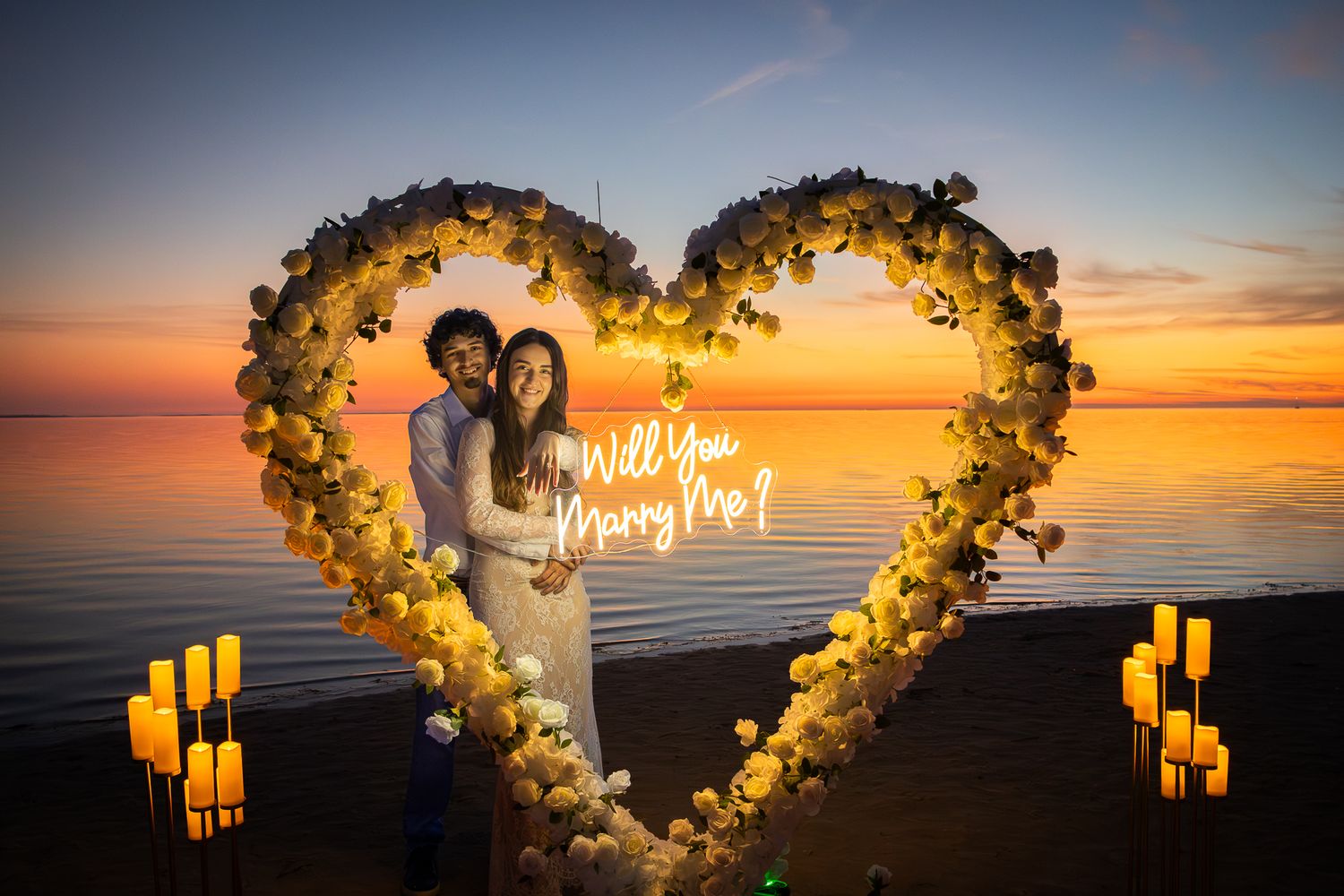 Couple standing inside floral heart-shaped arch with Will You Marry Me sign at sunset by the water.