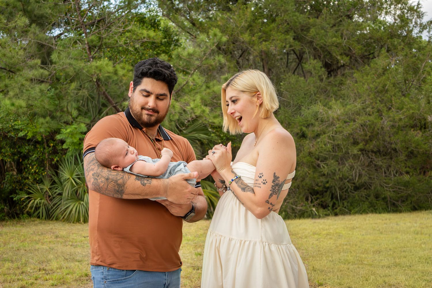 Young couple with tattoos smiling and holding newborn baby together in green outdoor park setting.