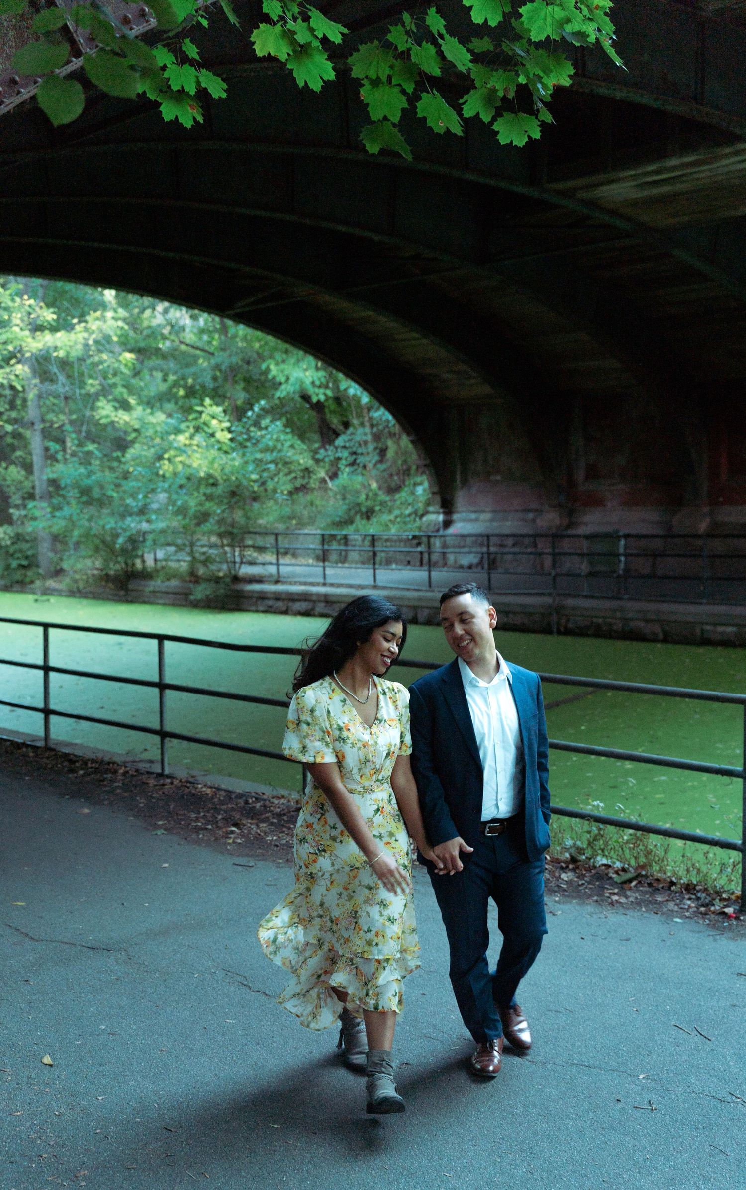 A couple walks hand-in-hand under a stone bridge in a lush park, woman in floral dress, man in navy suit.
