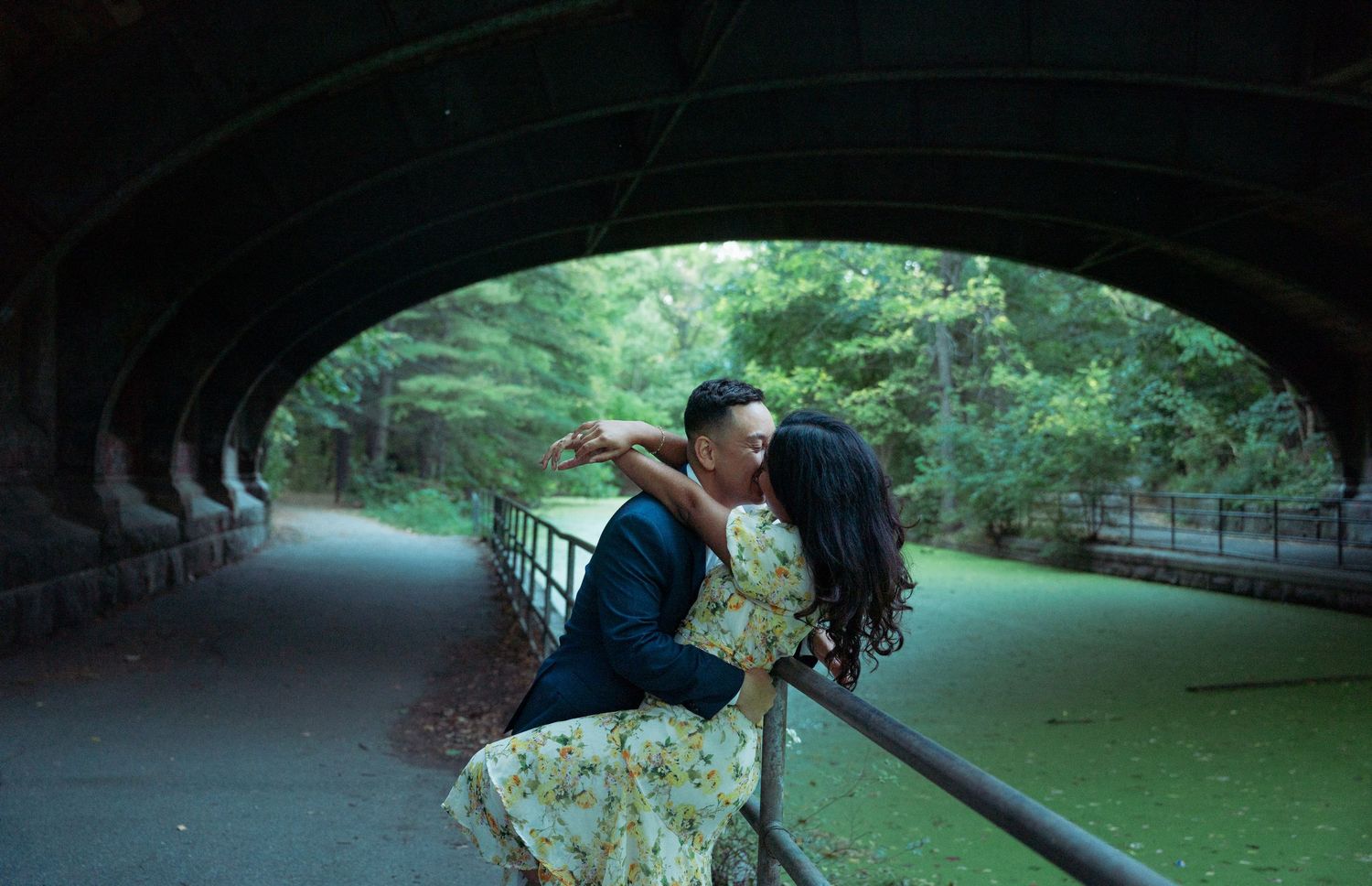 Couple sharing a romantic kiss under a stone bridge arch surrounded by lush green trees in a park.