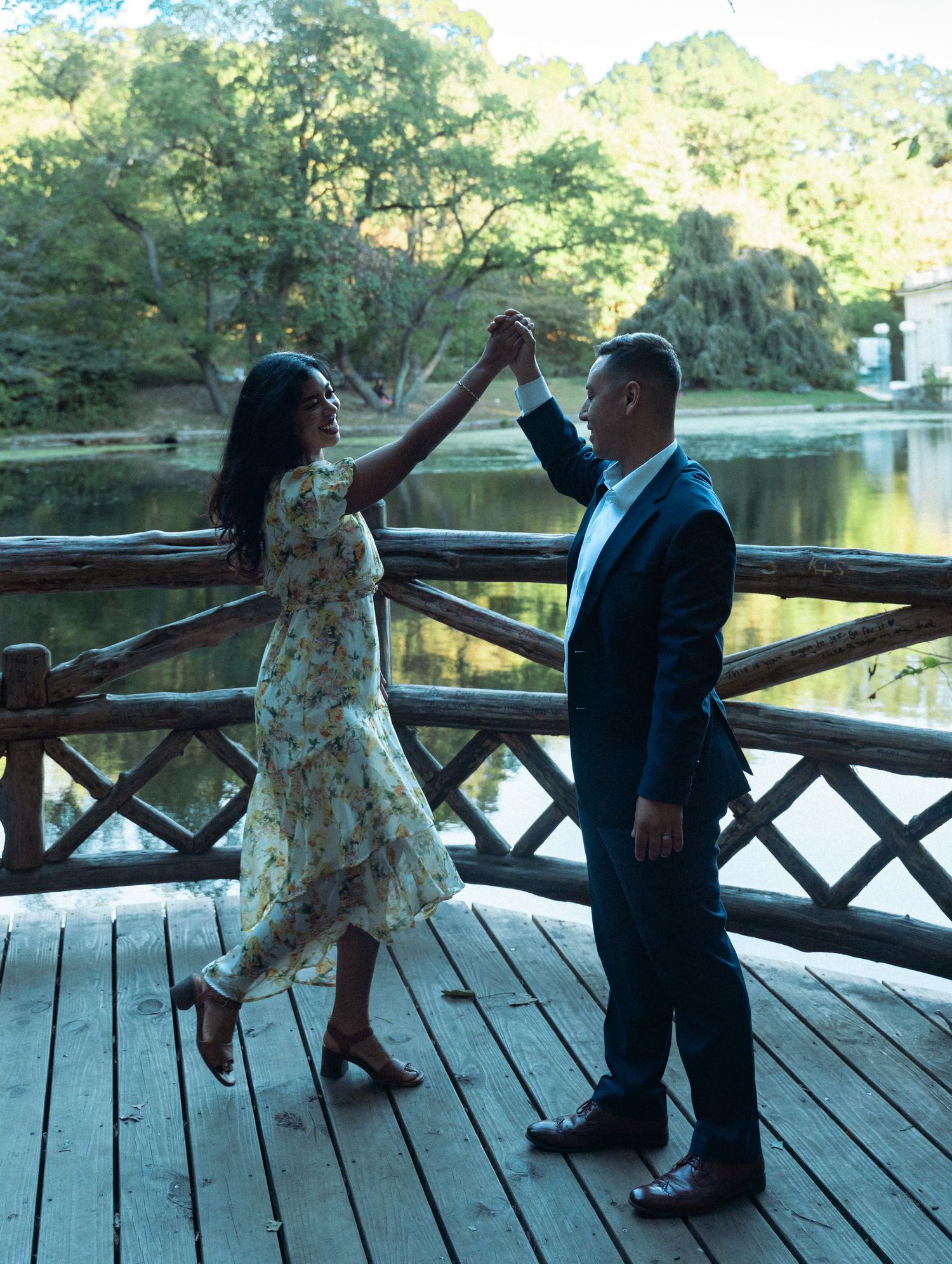 Couple dancing on a wooden deck by a serene lake, woman in floral dress, man in navy suit, surrounded by lush trees.