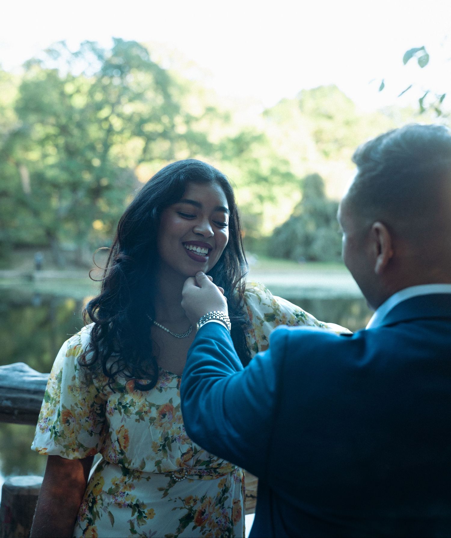 A man in a blue suit tenderly touches a smiling woman's chin outdoors near a serene pond surrounded by trees.