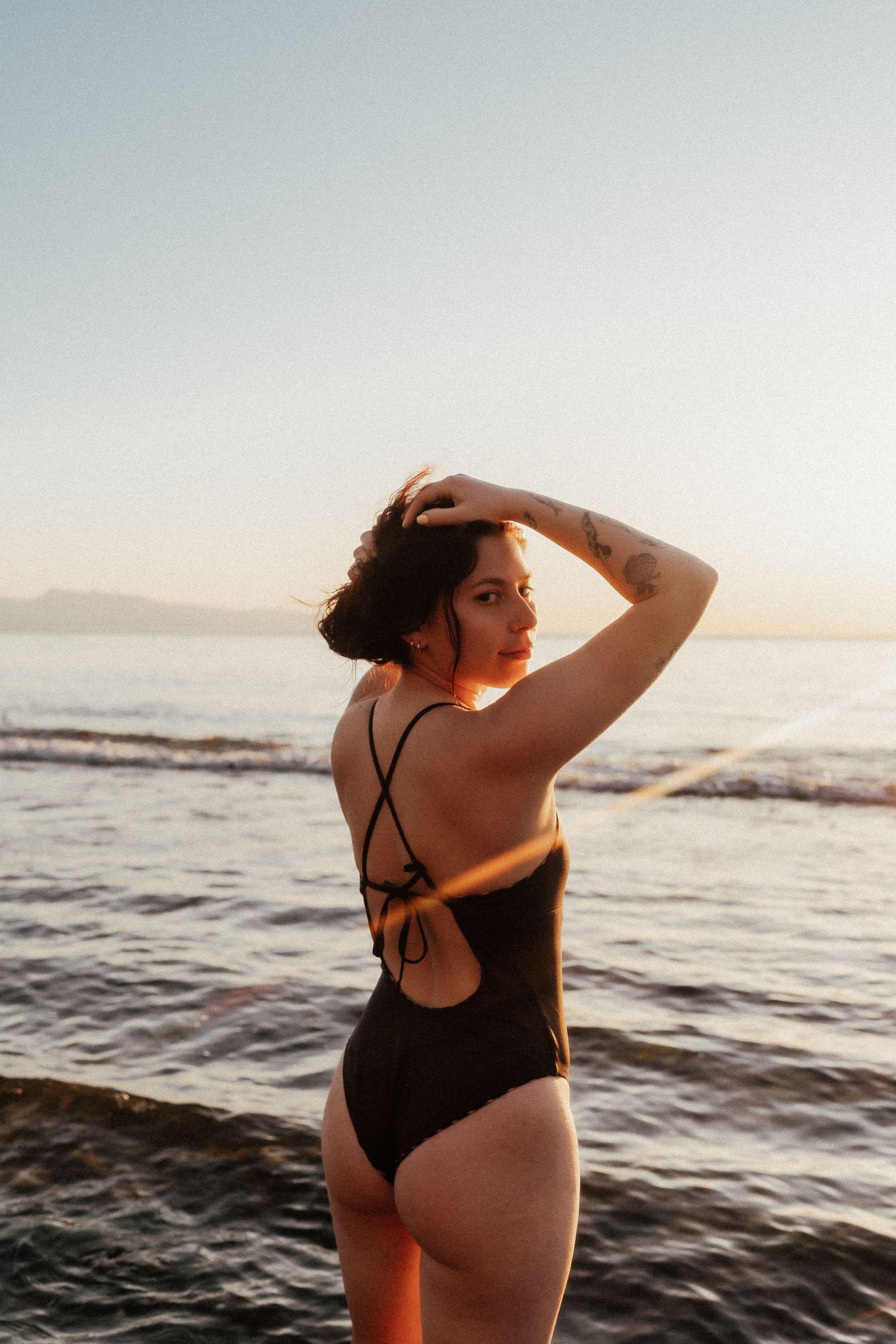 Woman in black swimsuit poses by the ocean at sunset, looking back over her shoulder with golden light.